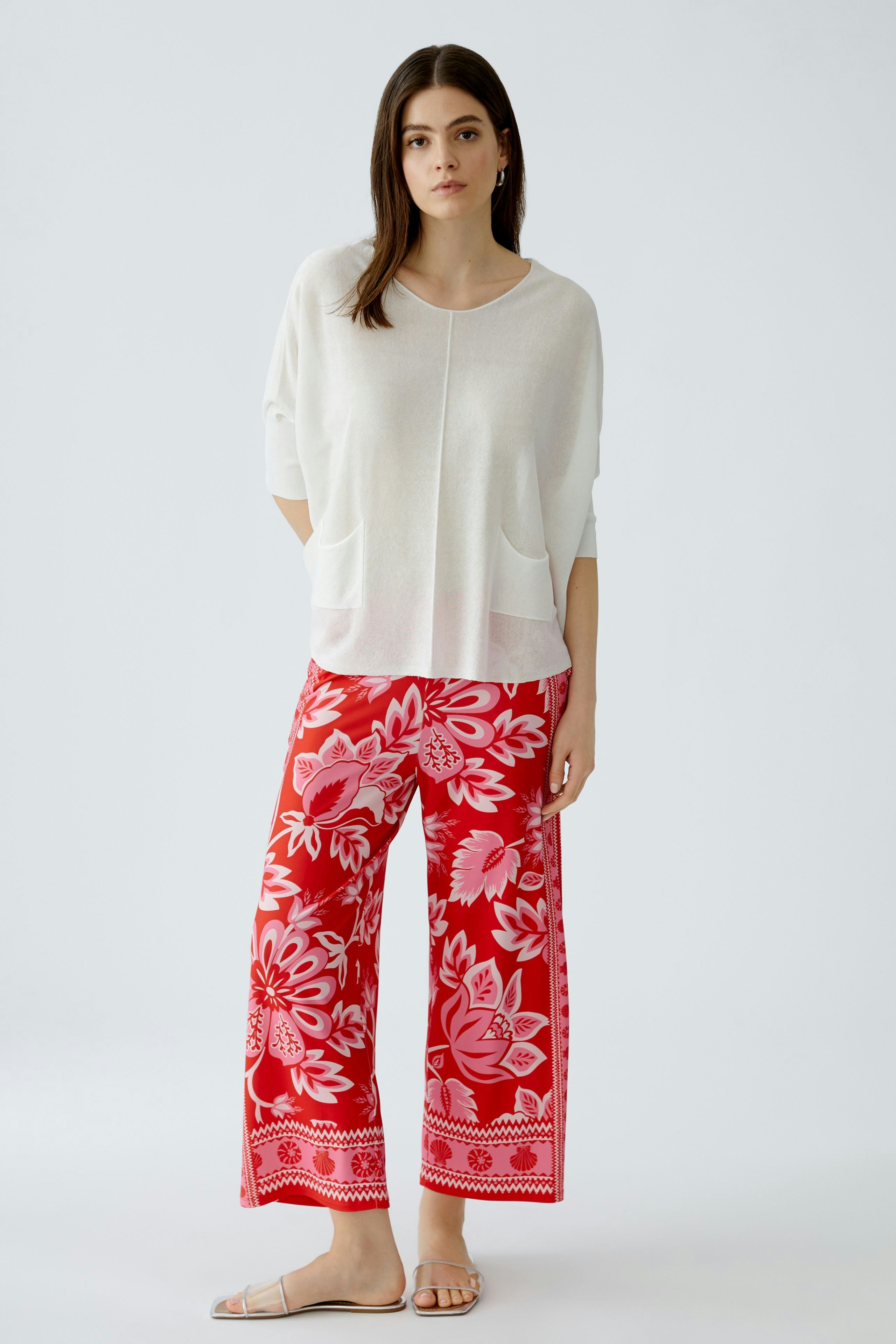 Lässiger Damen-Pullover in Weiß, kombiniert mit bunten, floralen Hosen und Sandalen.