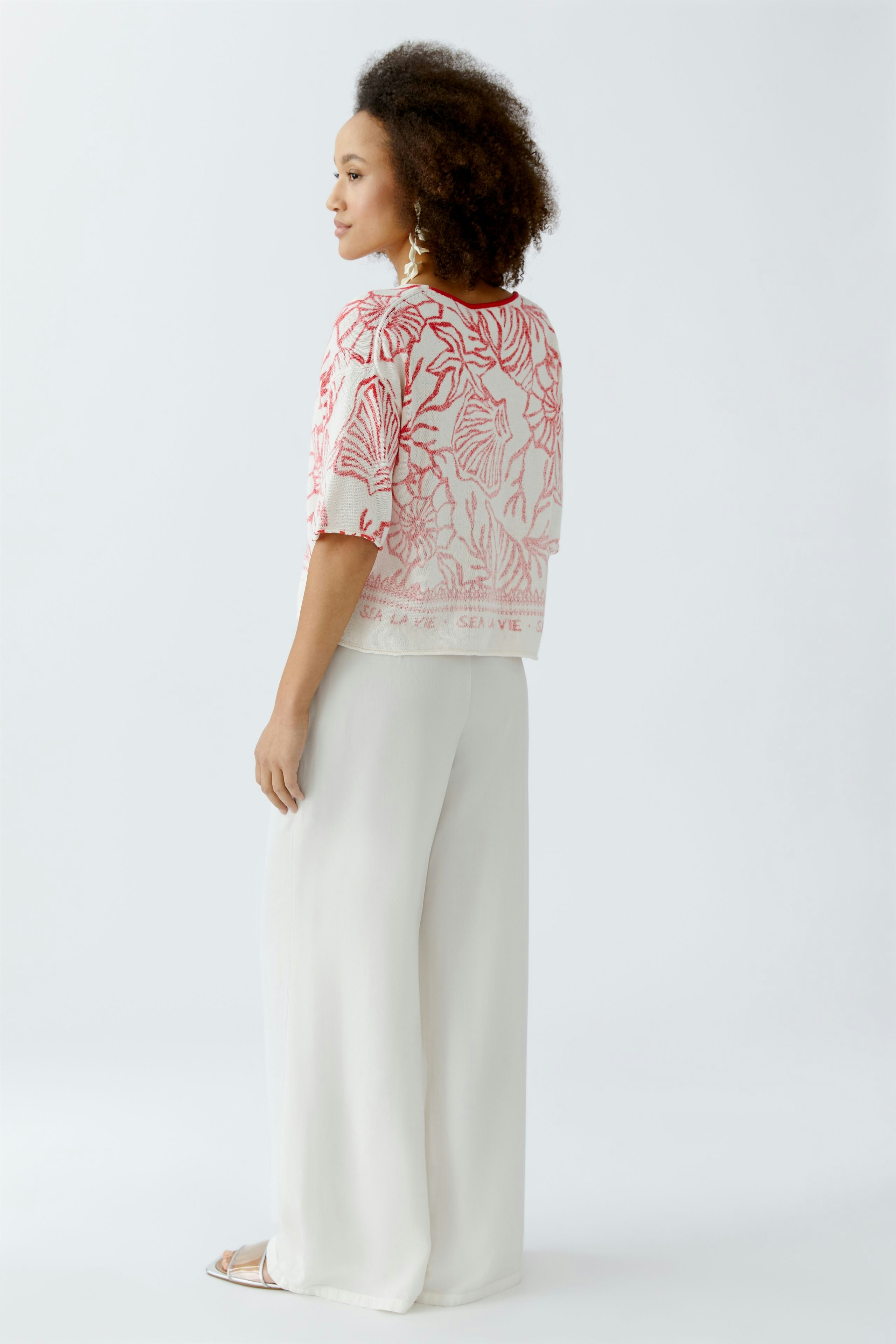 Damen-Pullover: Frau in einem roten, floral gemusterten Pullover mit lockerer Passform und weißen Hosen.