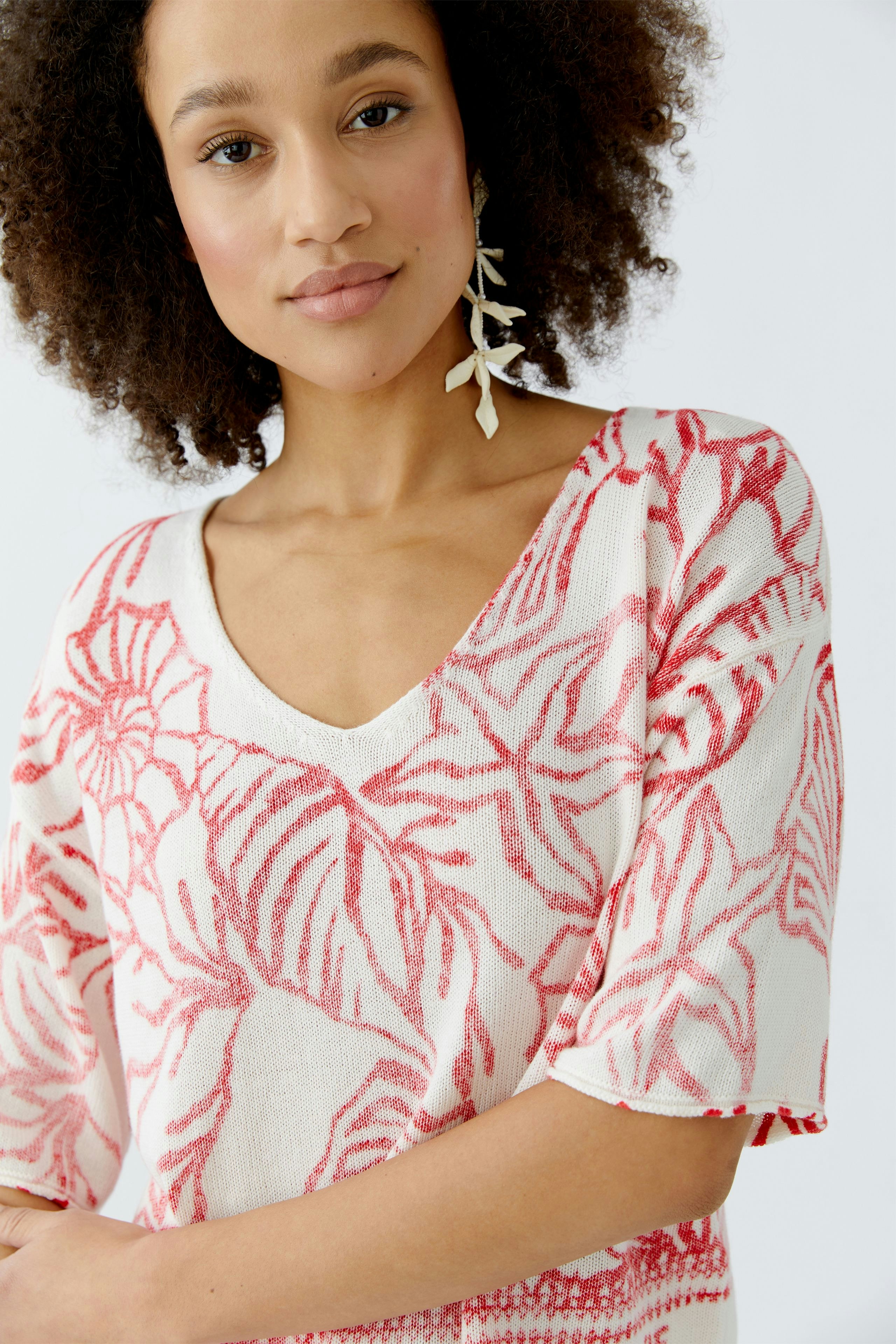 Damen-Pullover: Frau mit lockigem Haar trägt einen hellen Pullover mit floralem Muster in Rot.