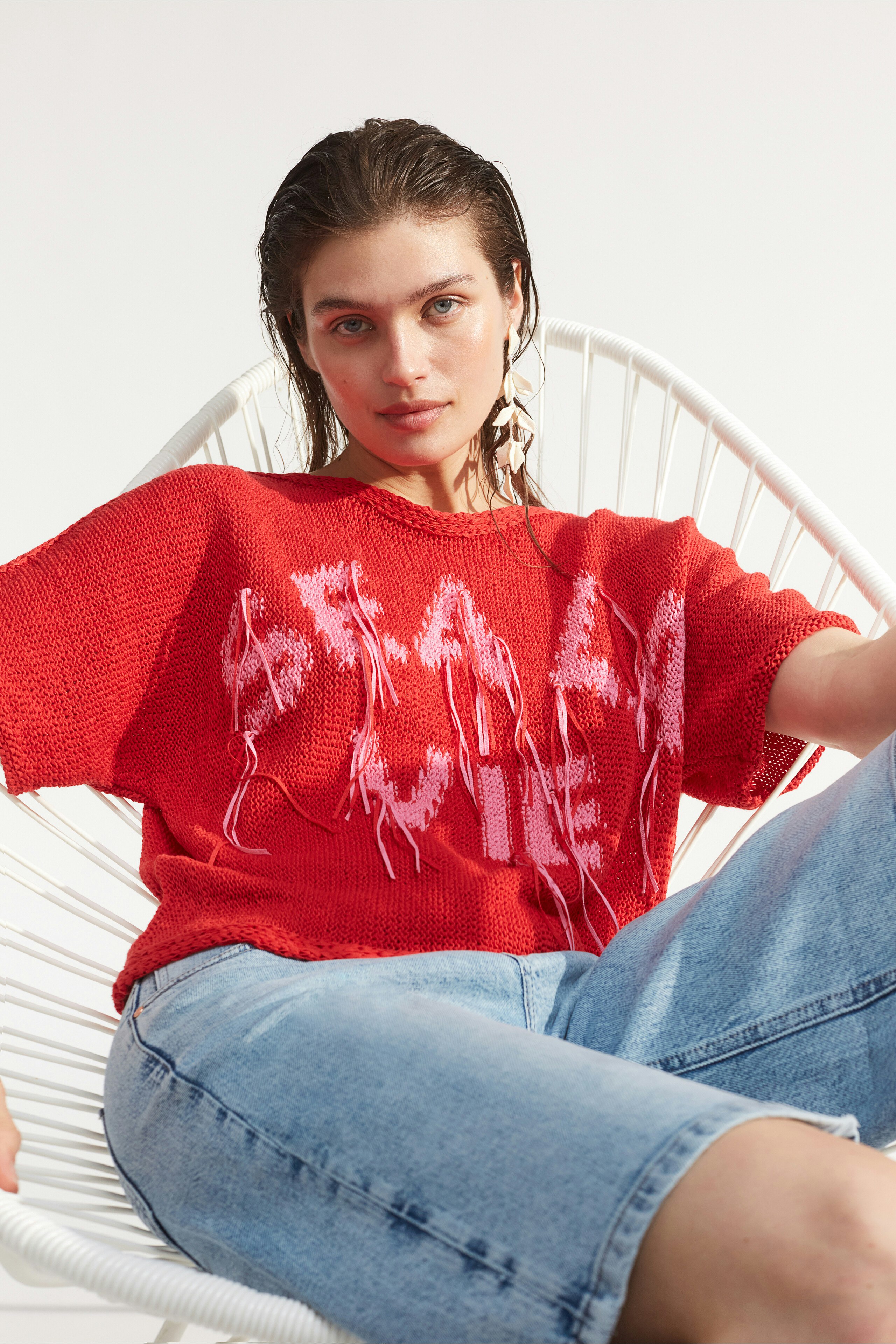 Casual women's pullover: Young woman in a red knit pullover with graphic print, relaxed in a chair.