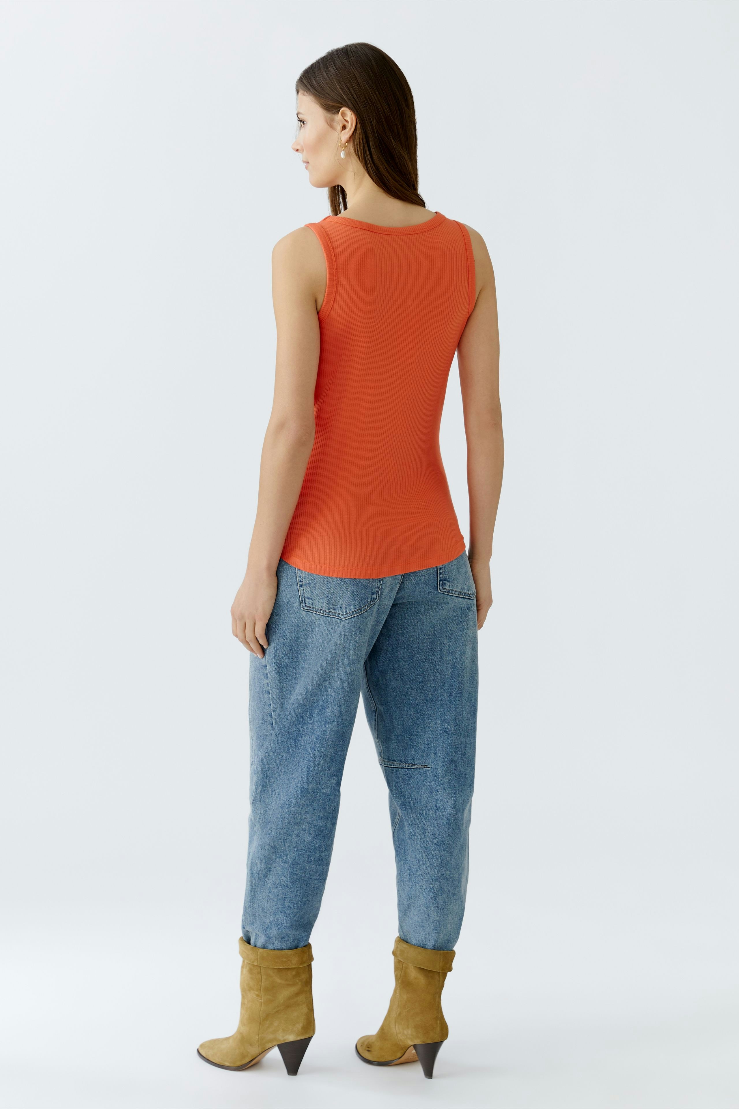 Bequeme Damen-Top: Frau in orangefarbenem Tanktop und lässigen Jeans steht entspannt mit dem Rücken zur Kamera.