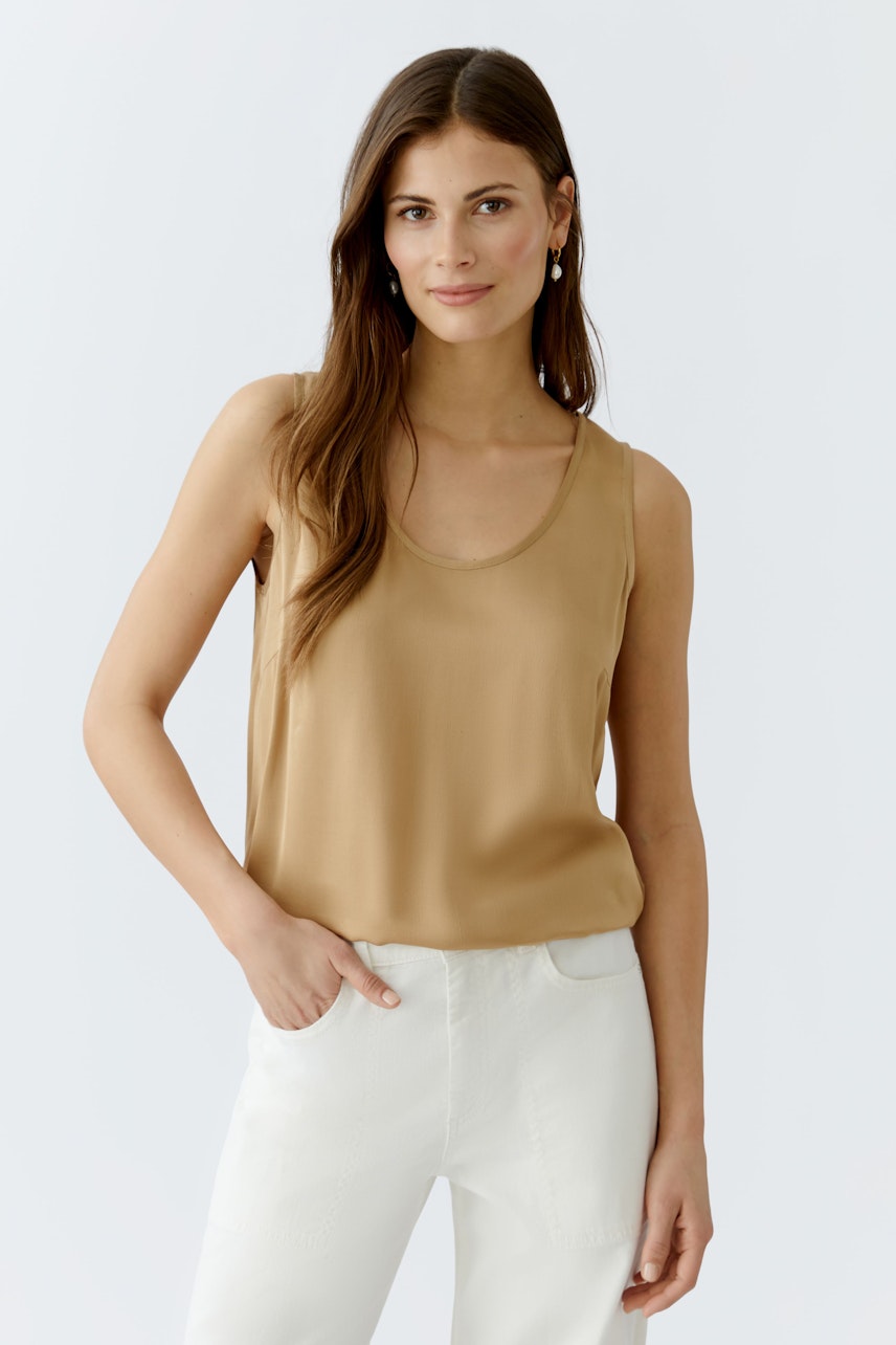 Beige Damen-Top: Frau trägt ein schlichtes, ärmelloses Top in Beige, kombiniert mit hellen Hosen.