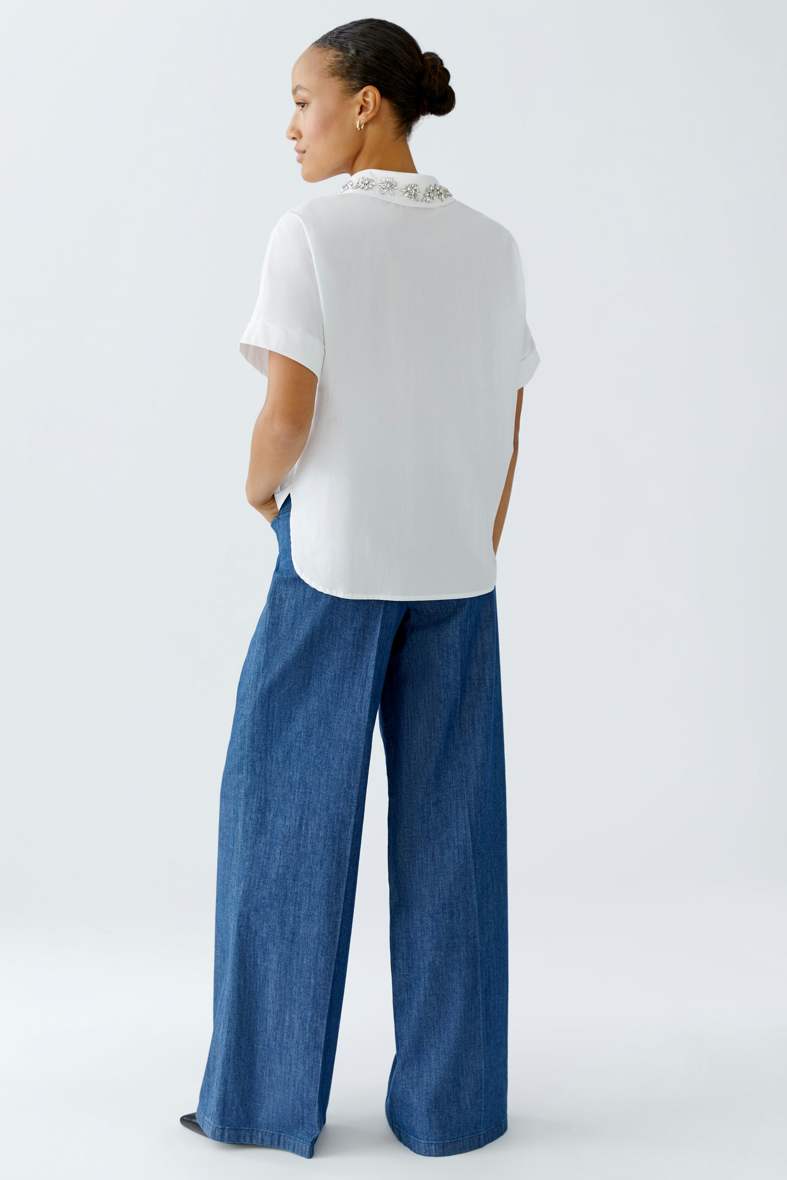 Damen-Bluse: Frau in weißer Bluse mit besticktem Kragen und lässiger Jeanshose von hinten.
