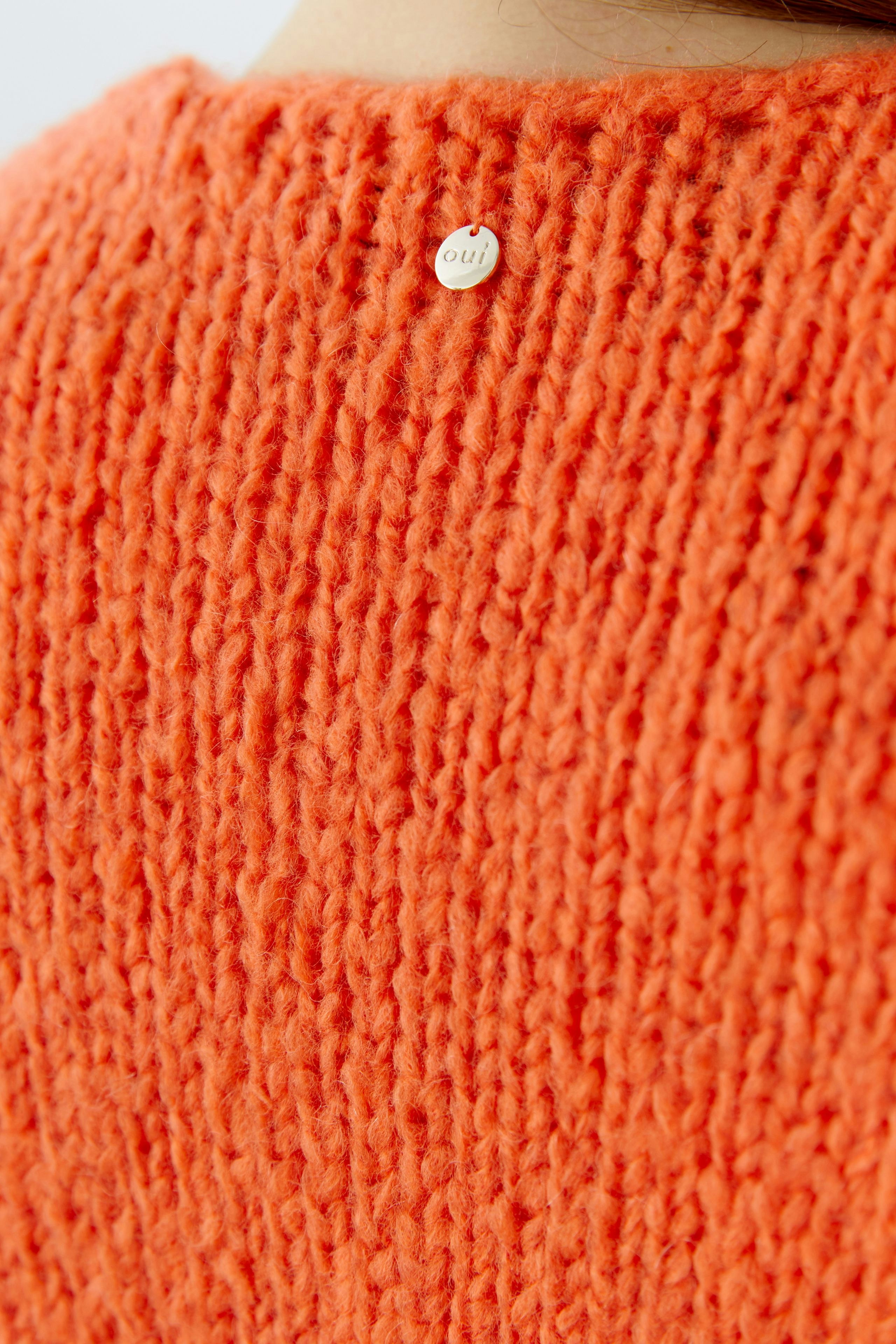 Strickjacke in leuchtendem Orange mit grober Struktur und Logo-Detail auf der Rückseite.