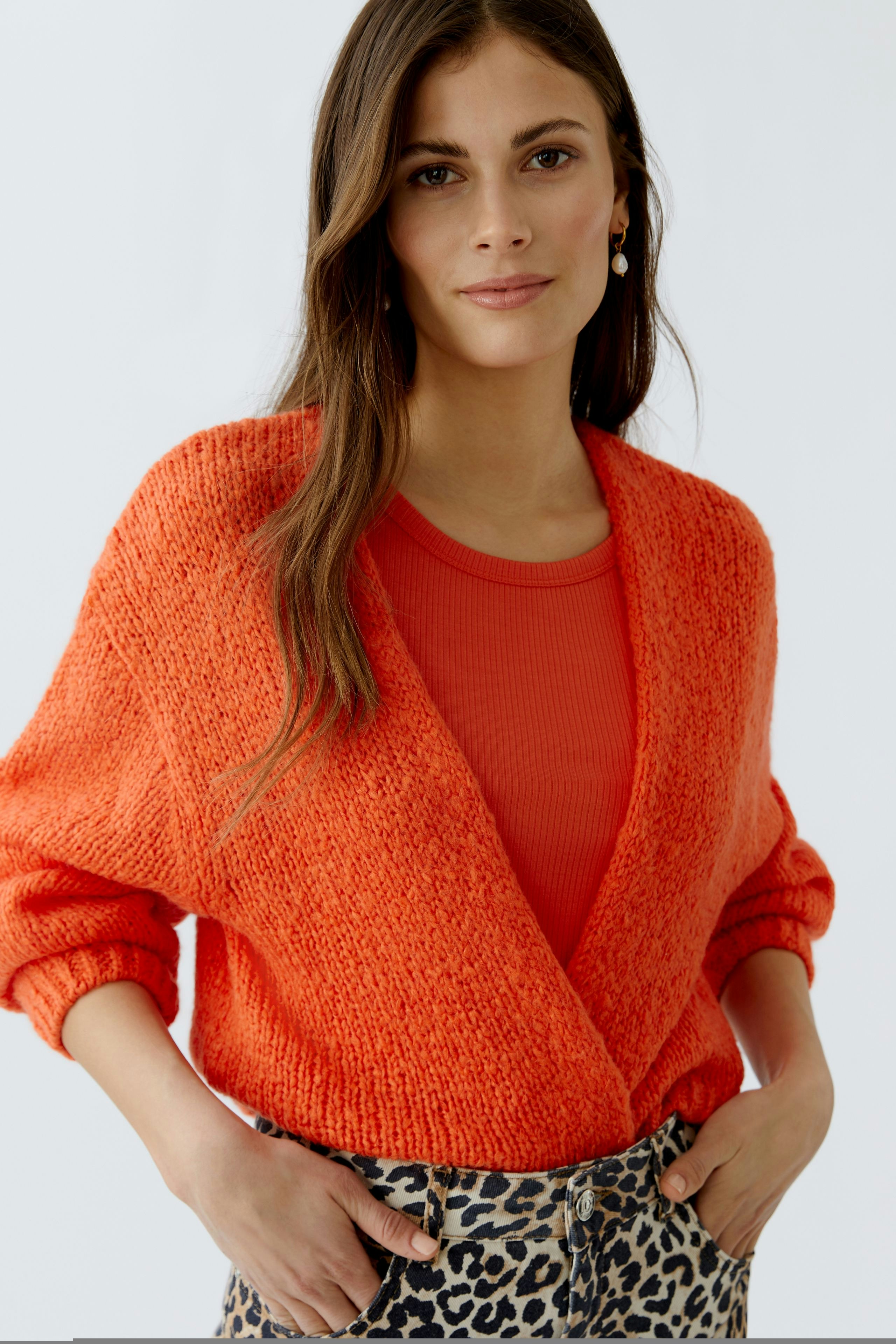 Lässige Damen-Strickjacke in leuchtendem Orange, kombiniert mit einem roten Oberteil und Jeans im Leo-Print.