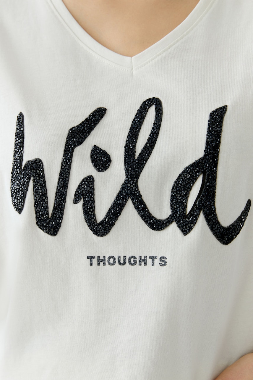 Bequemes Damen-T-Shirt: Frau trägt ein weißes Shirt mit glitzerndem Schriftzug 'Wild Thoughts'.