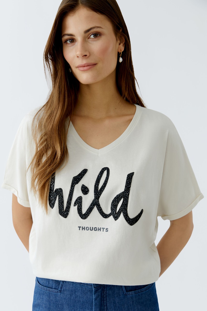 Bequeme Damen-T-Shirt: Frau trägt ein helles T-Shirt mit glitzerndem Schriftzug 'Wild Thoughts'.