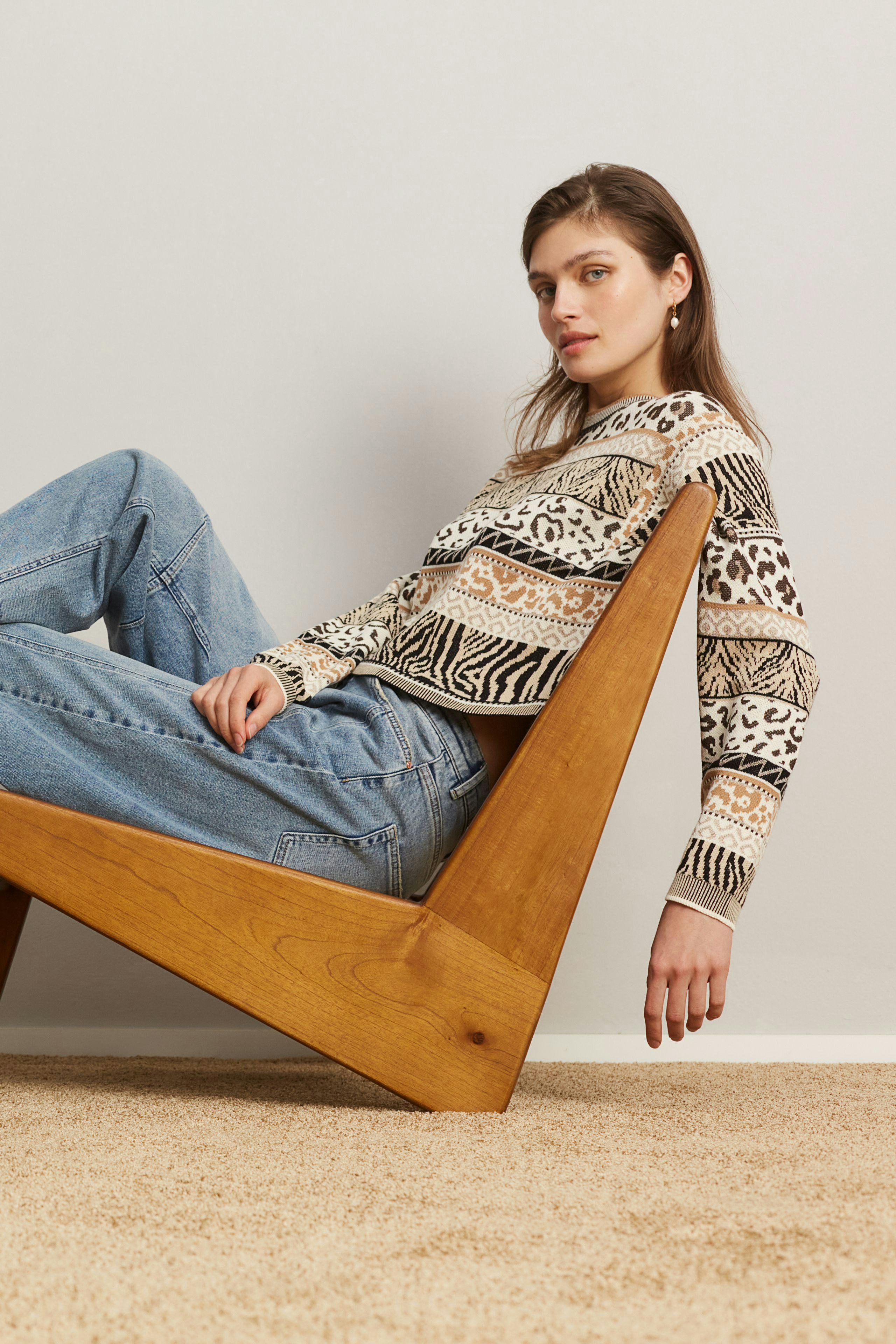 Damen-Pullover: Frau in gemustertem Pullover mit lässigen Jeans sitzt entspannt auf einem Holzstuhl.