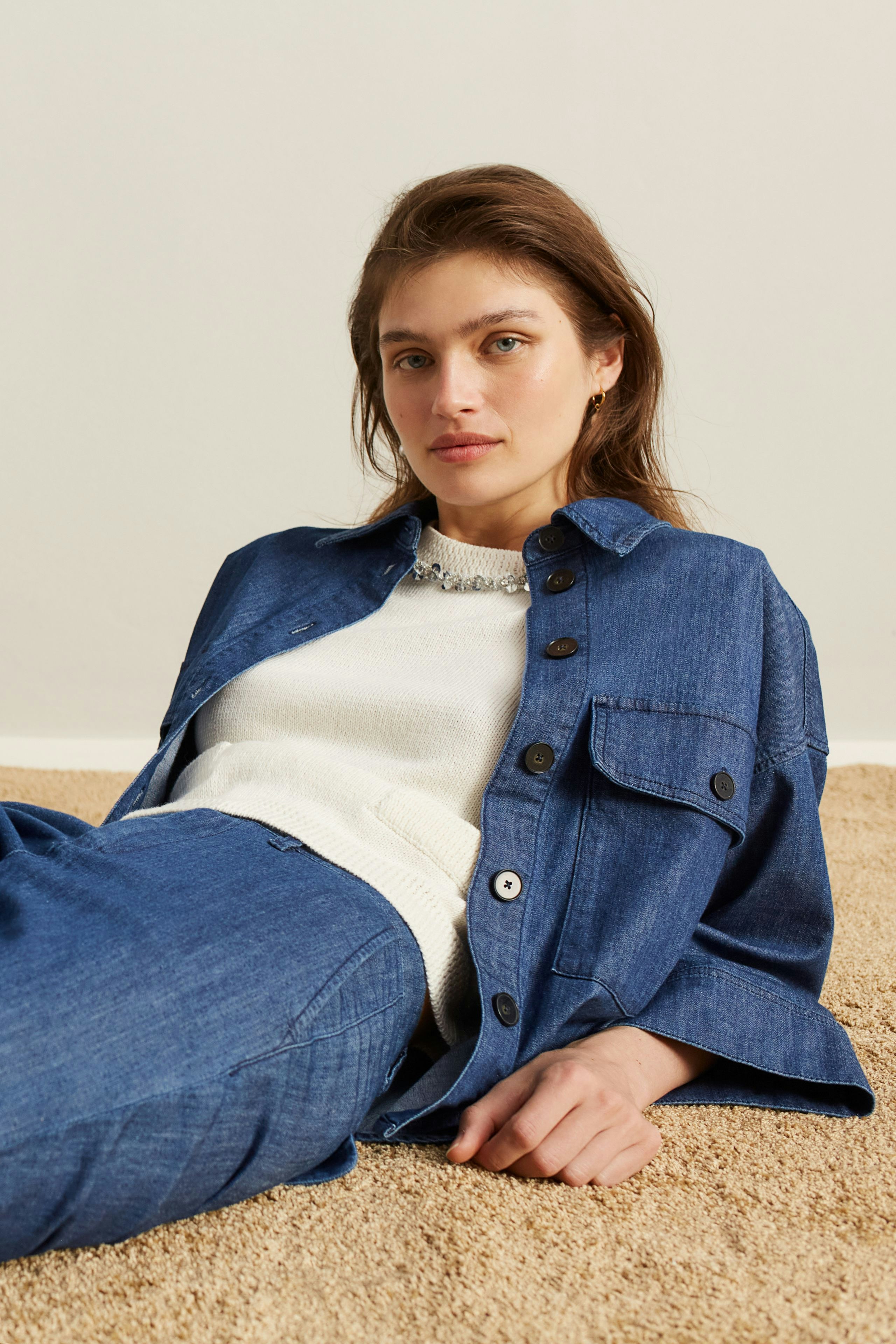 Damen-Pullover: Frau in hellem Strickpullover mit lässiger Jeansjacke sitzt entspannt auf dem Boden.