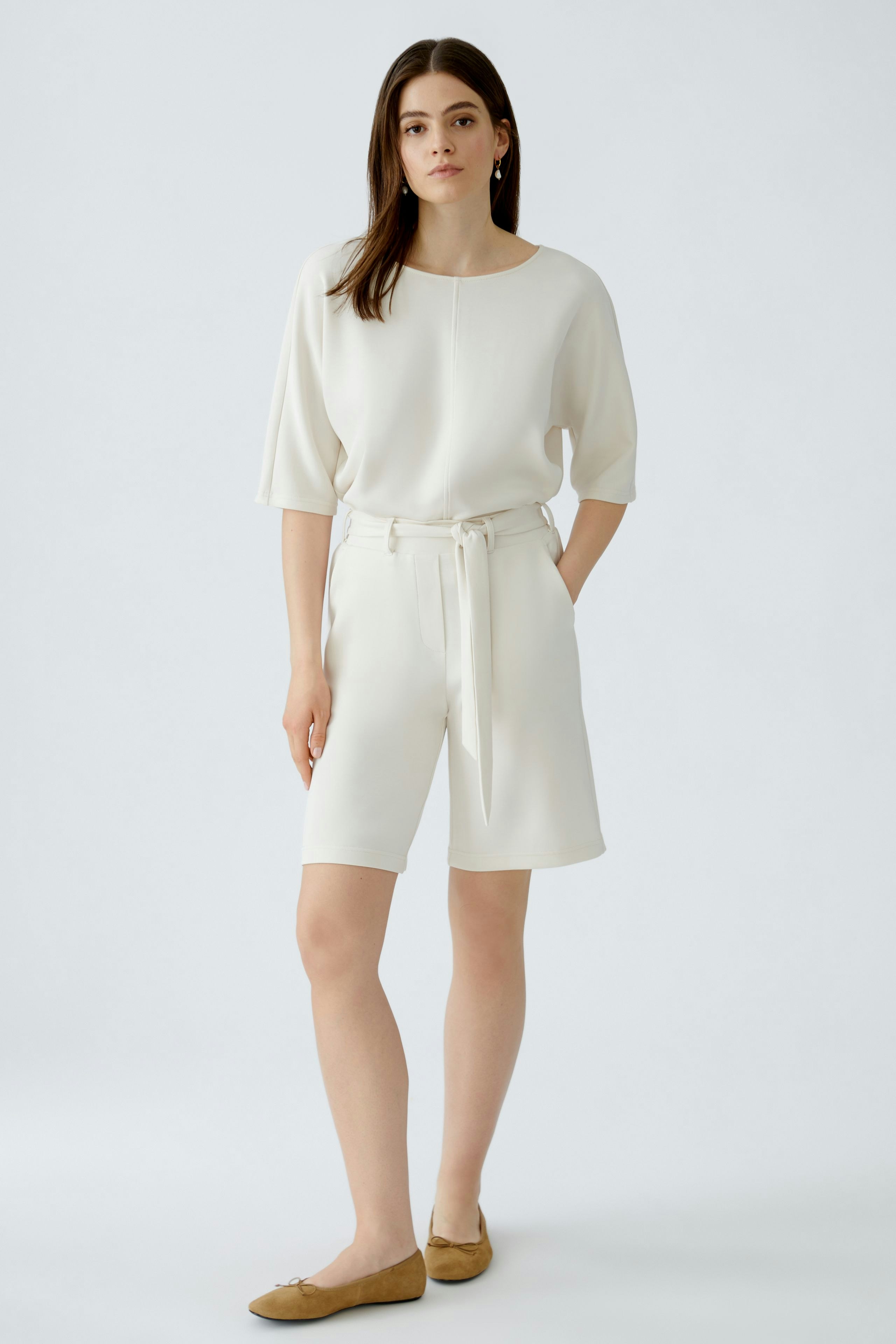Bequeme Damen-Bermuda: Junge Frau in weißen Shorts mit Gürtel und lässigem Oberteil steht entspannt.