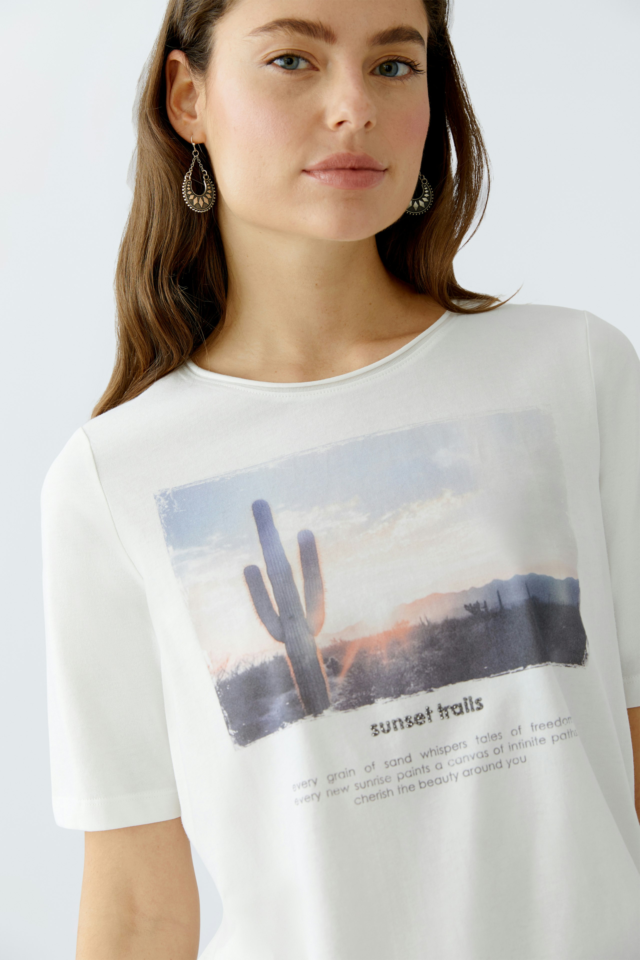 Damen-T-Shirt: Frau trägt ein weißes T-Shirt mit einem bunten Wüstenmotiv und lässigem Schnitt.