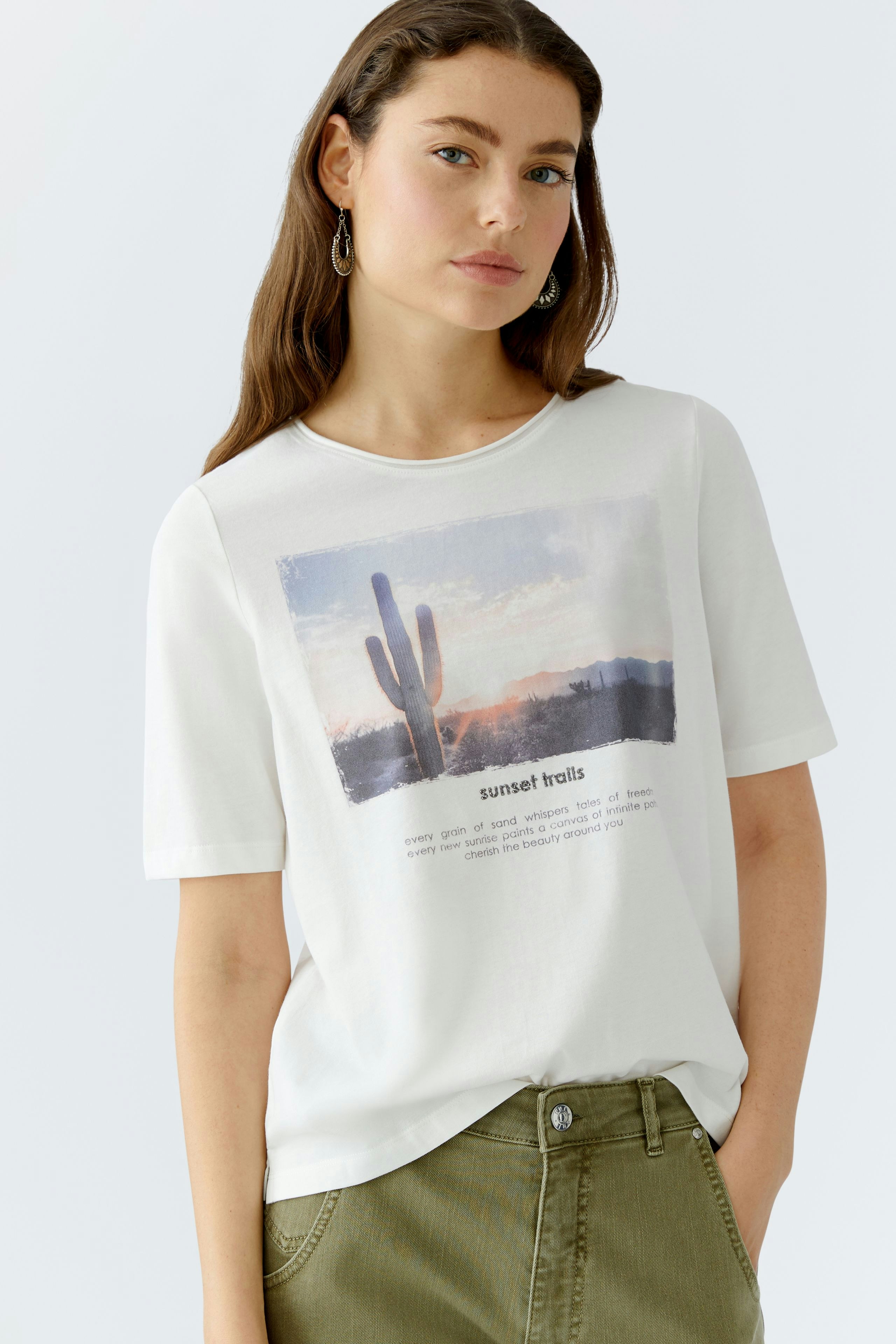 Damen-T-Shirt: Junge Frau trägt ein weißes T-Shirt mit einem bunten Wüstenmotiv und lässigen Hosen.