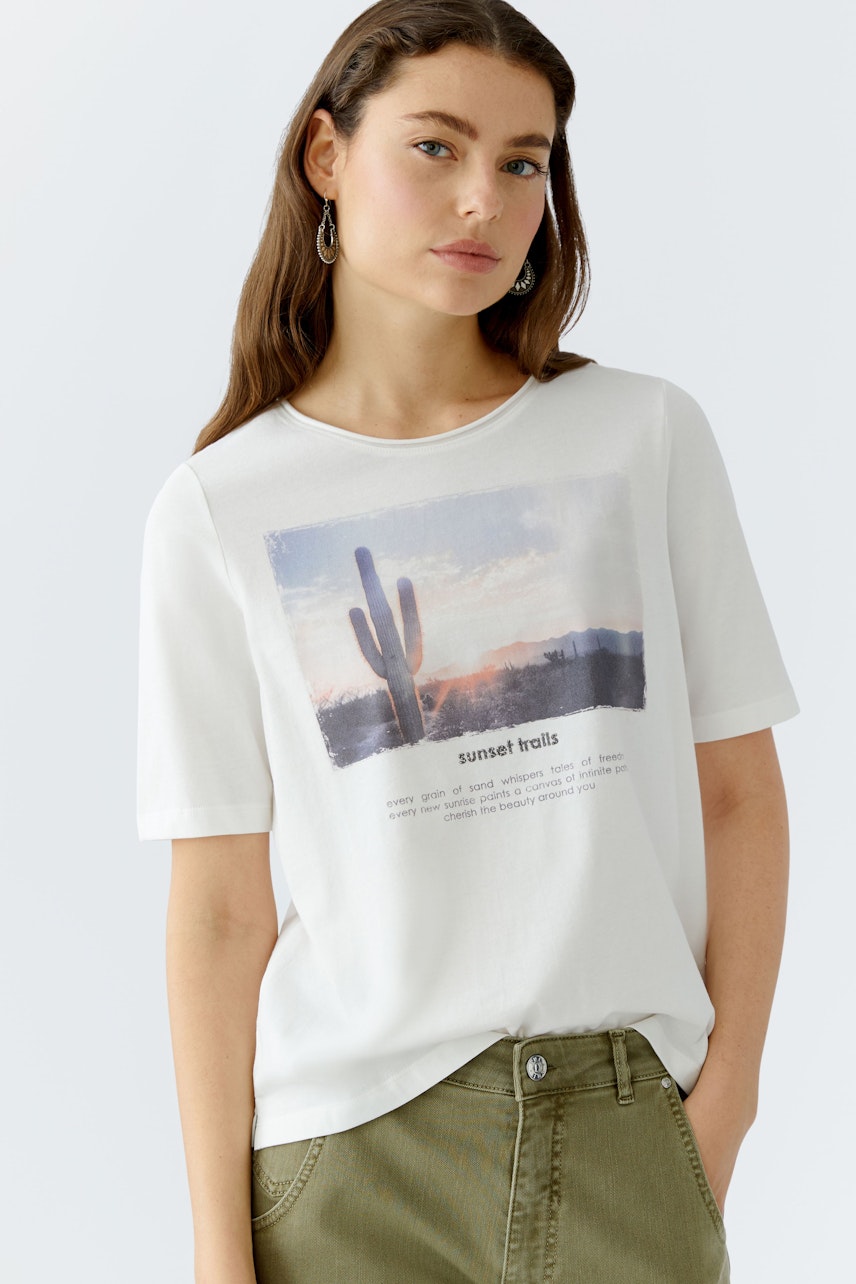 Damen-T-Shirt: Junge Frau trägt ein weißes T-Shirt mit einem bunten Wüstenmotiv und lässigen Hosen.