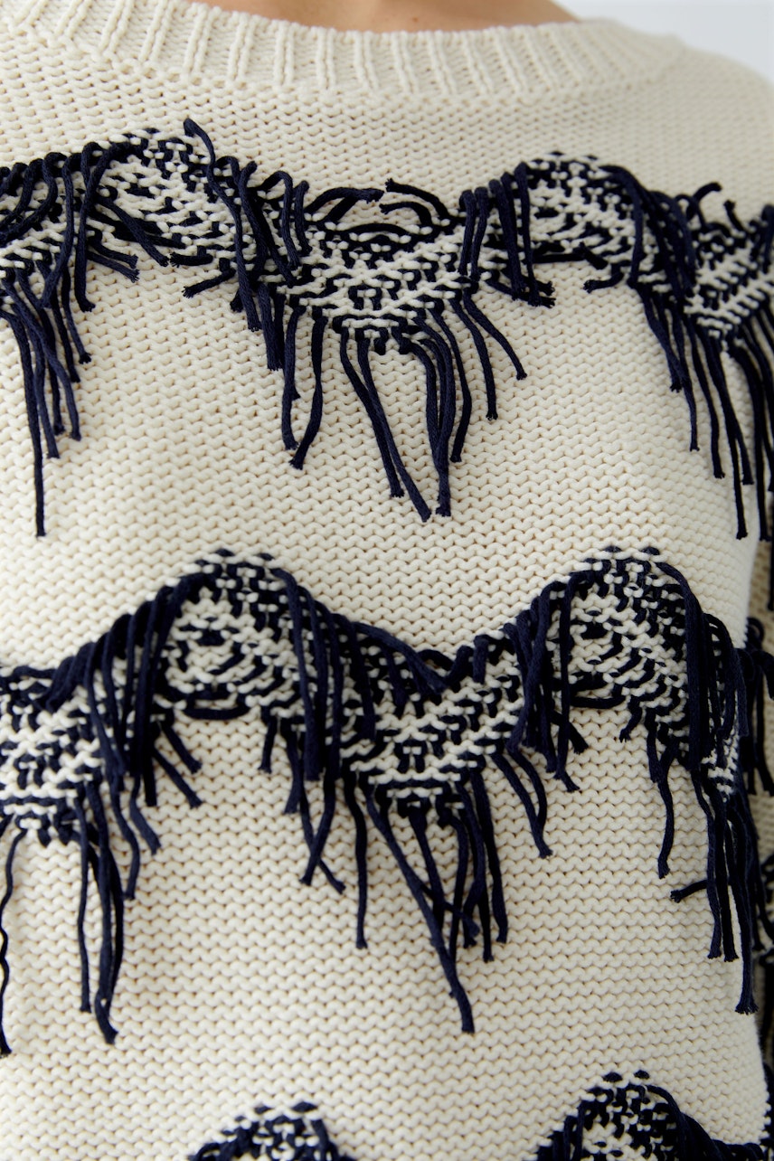 Casual women's pullover: Close-up of a cream pullover with navy fringes and textured patterns.