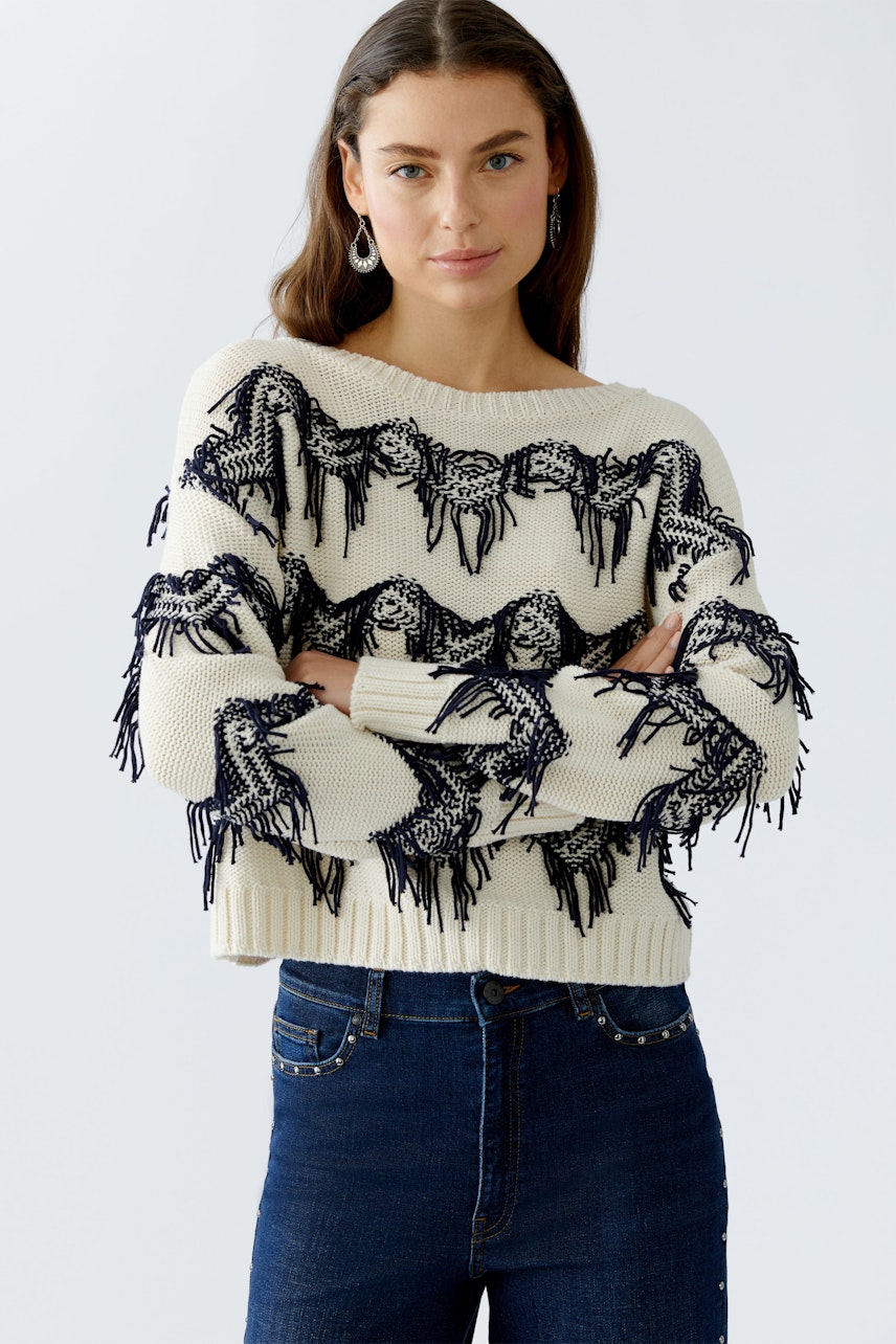 Casual women's pullover: Young woman in a cream pullover with navy fringe detailing, arms crossed.