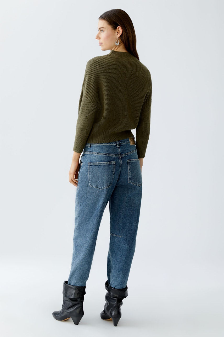 Damen-Pullover: Frau in olivgrünem Strickpullover mit lässigem Schnitt und Jeans im Hintergrund.