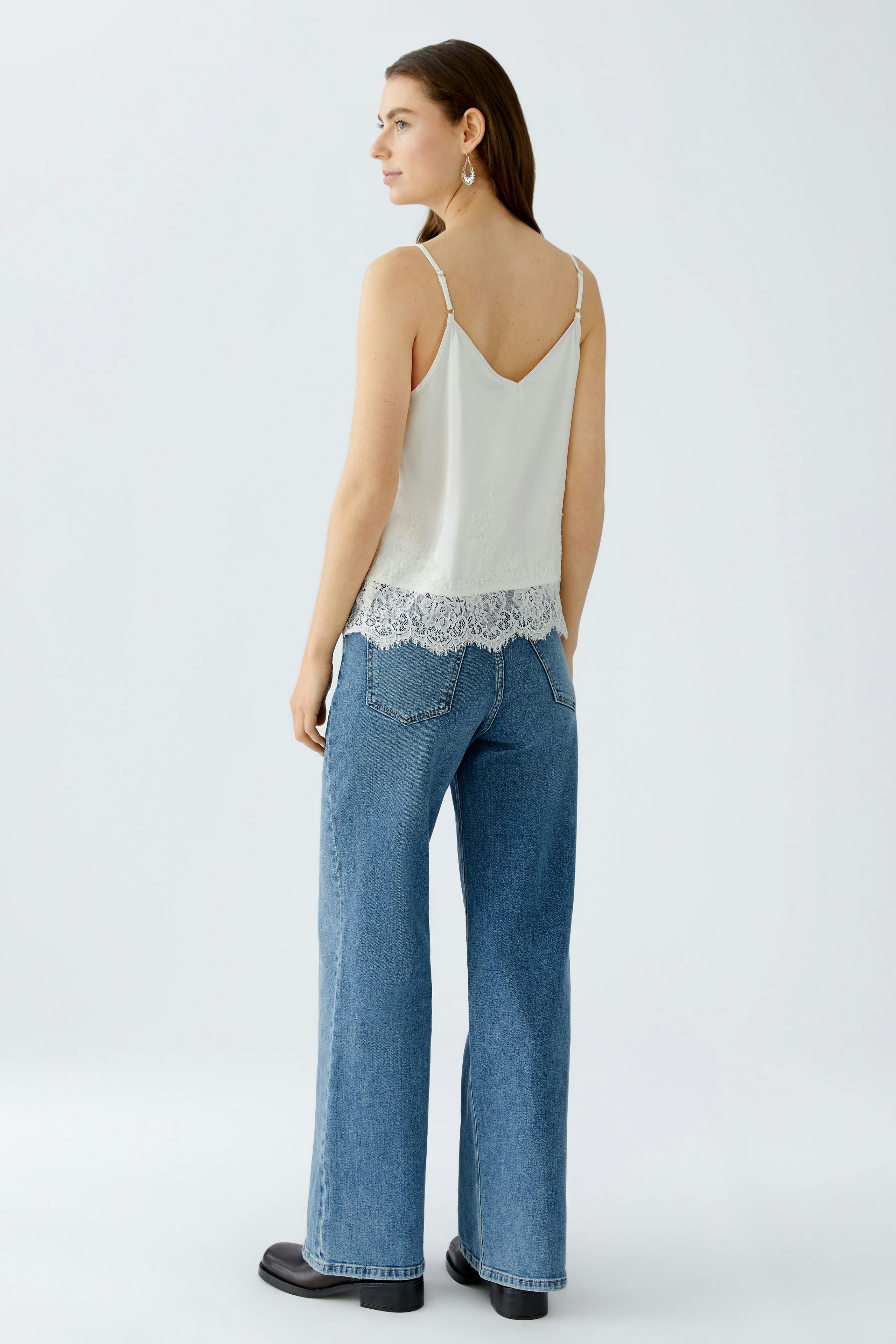 Casual women's top: Young woman in a light tank top with lace trim and wide jeans, standing confidently.