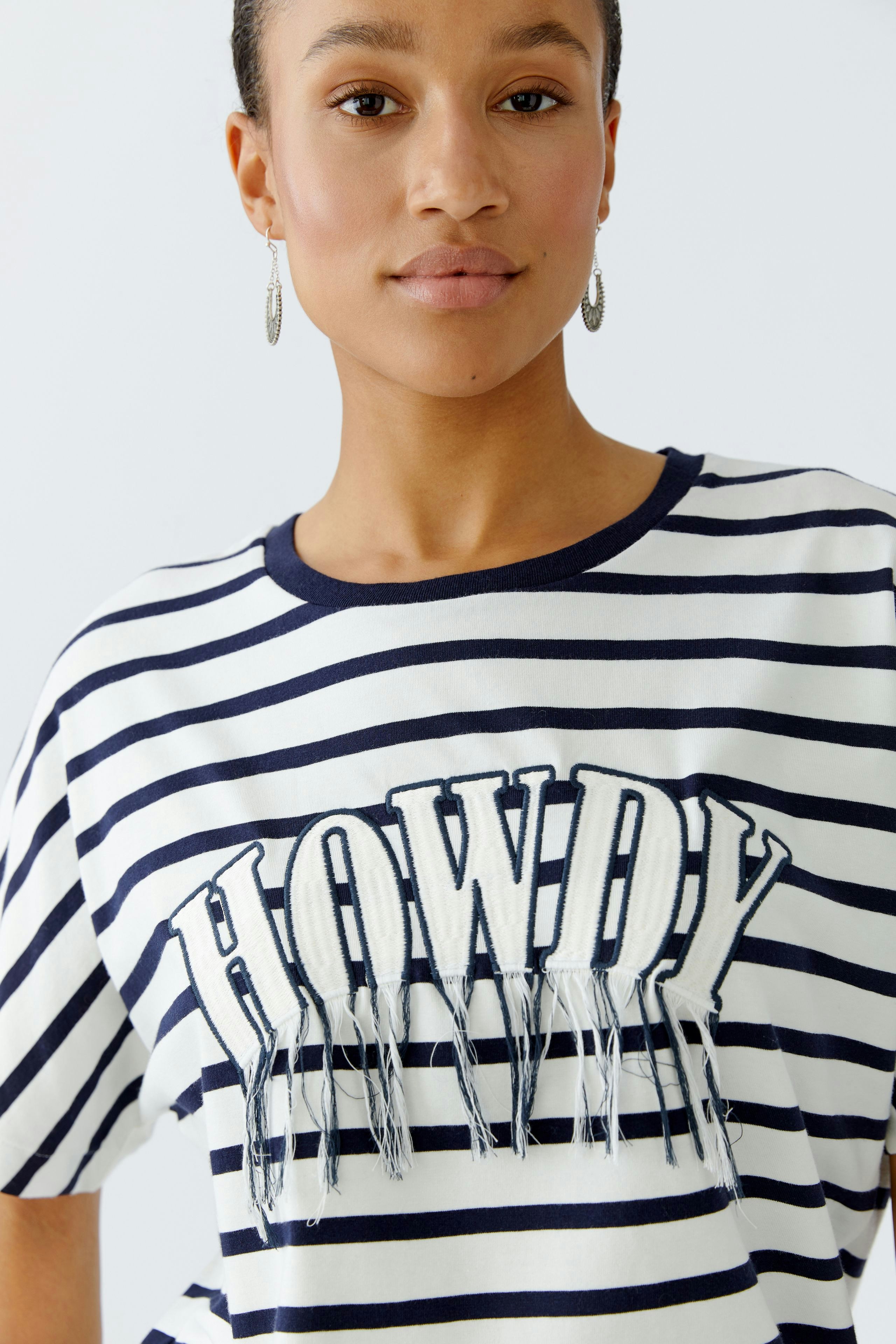 Casual women's T-shirt: A woman in a striped tee with 'HOWDY' print and fringes, smiling confidently.