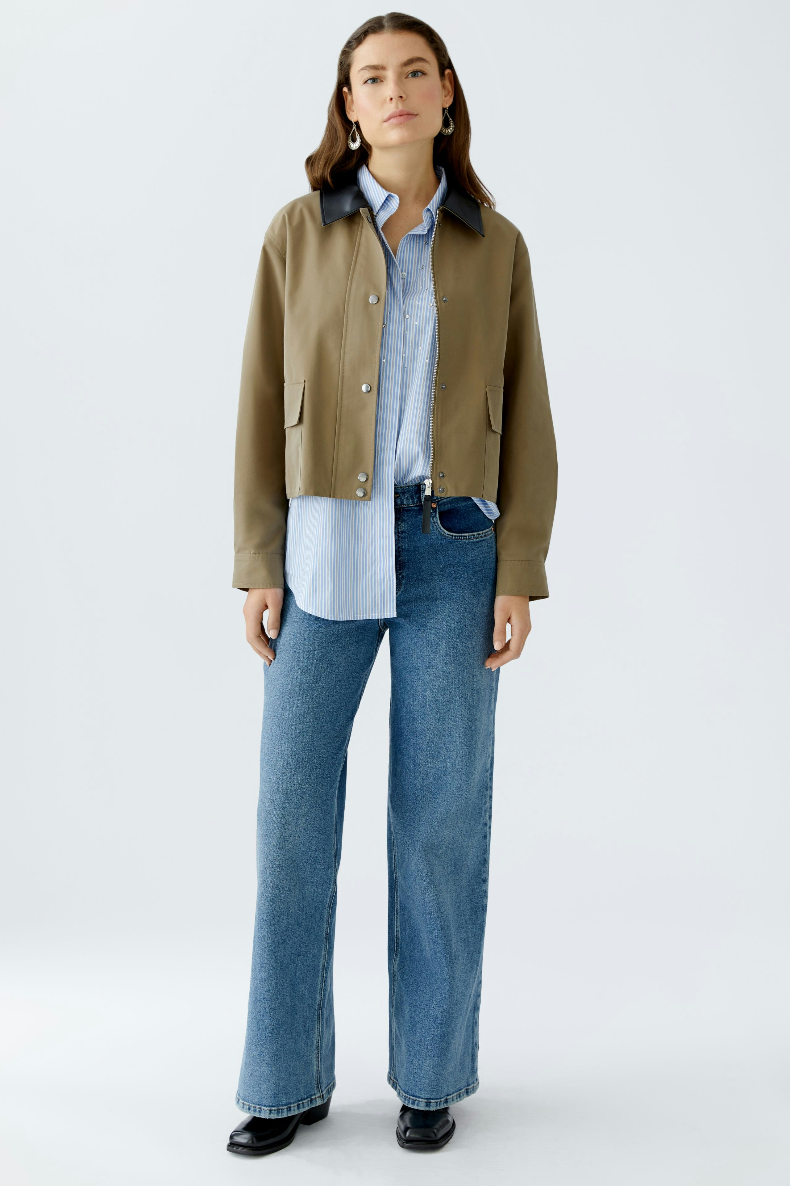 Moderne Damen-Jacke: Frau in beiger Jacke mit blauer Bluse und Jeans steht lässig vor neutralem Hintergrund.