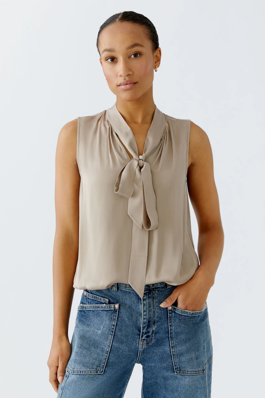 Sleeveless blouse: Woman in a beige blouse with a bow detail, paired with casual denim jeans.
