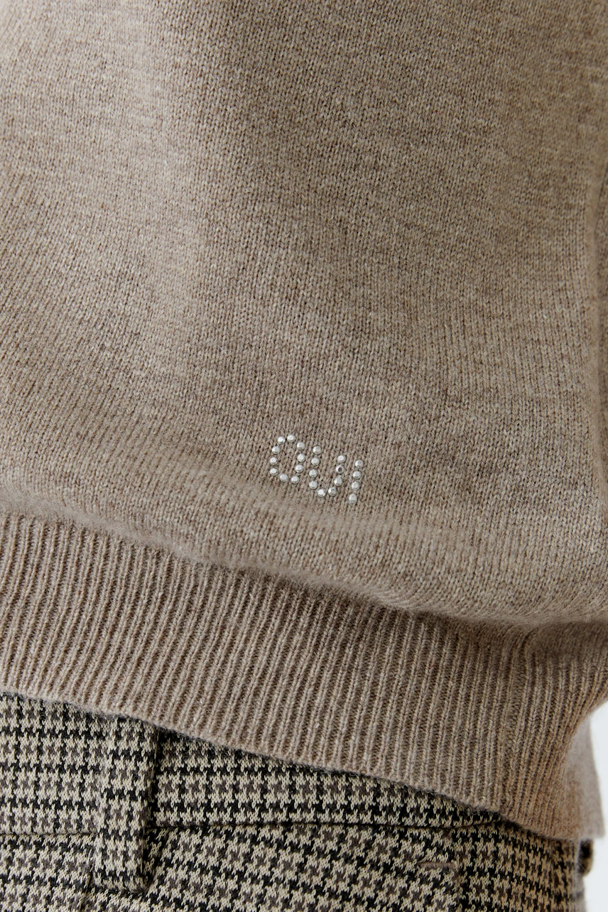 Beiger Damen-Pullover mit glitzerndem Logo, lässig kombiniert mit karierten Hosen.