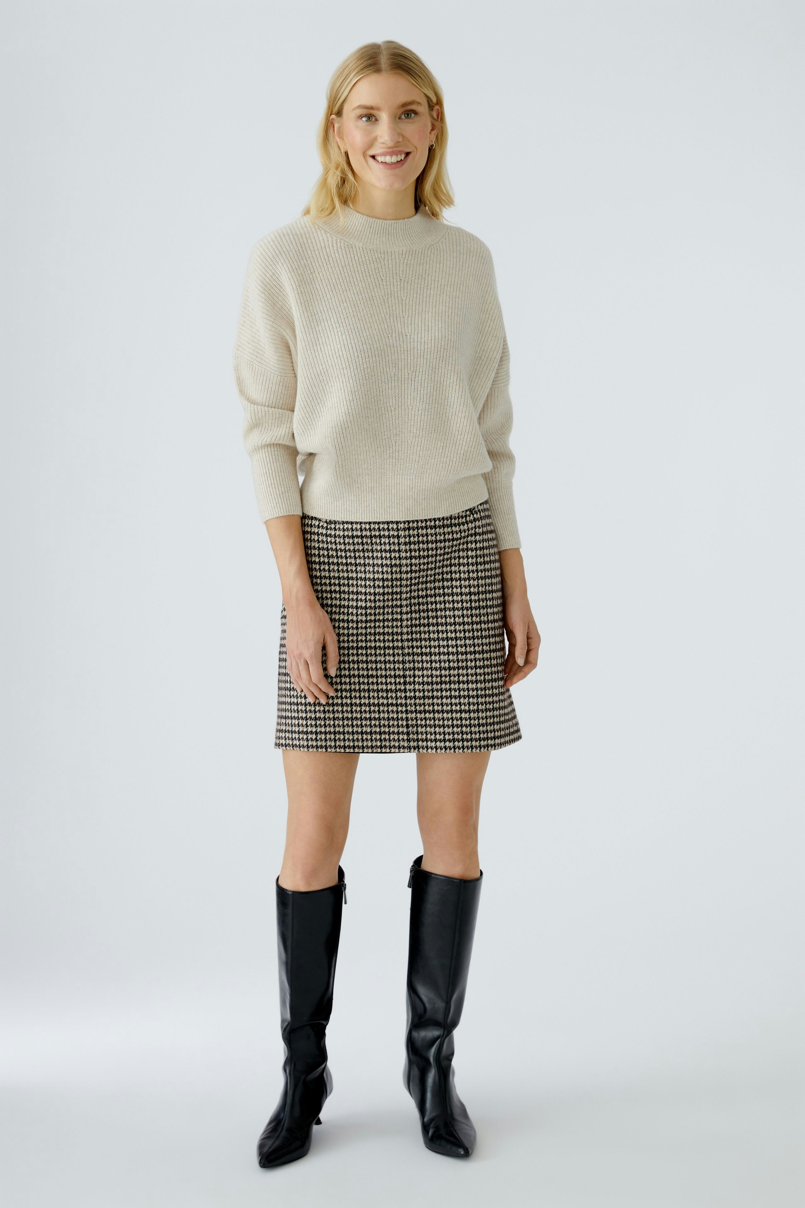 Damen-Pullover: Frau trägt einen beigen, lässigen Pullover zu einem karierten Rock und schwarzen Stiefeln.