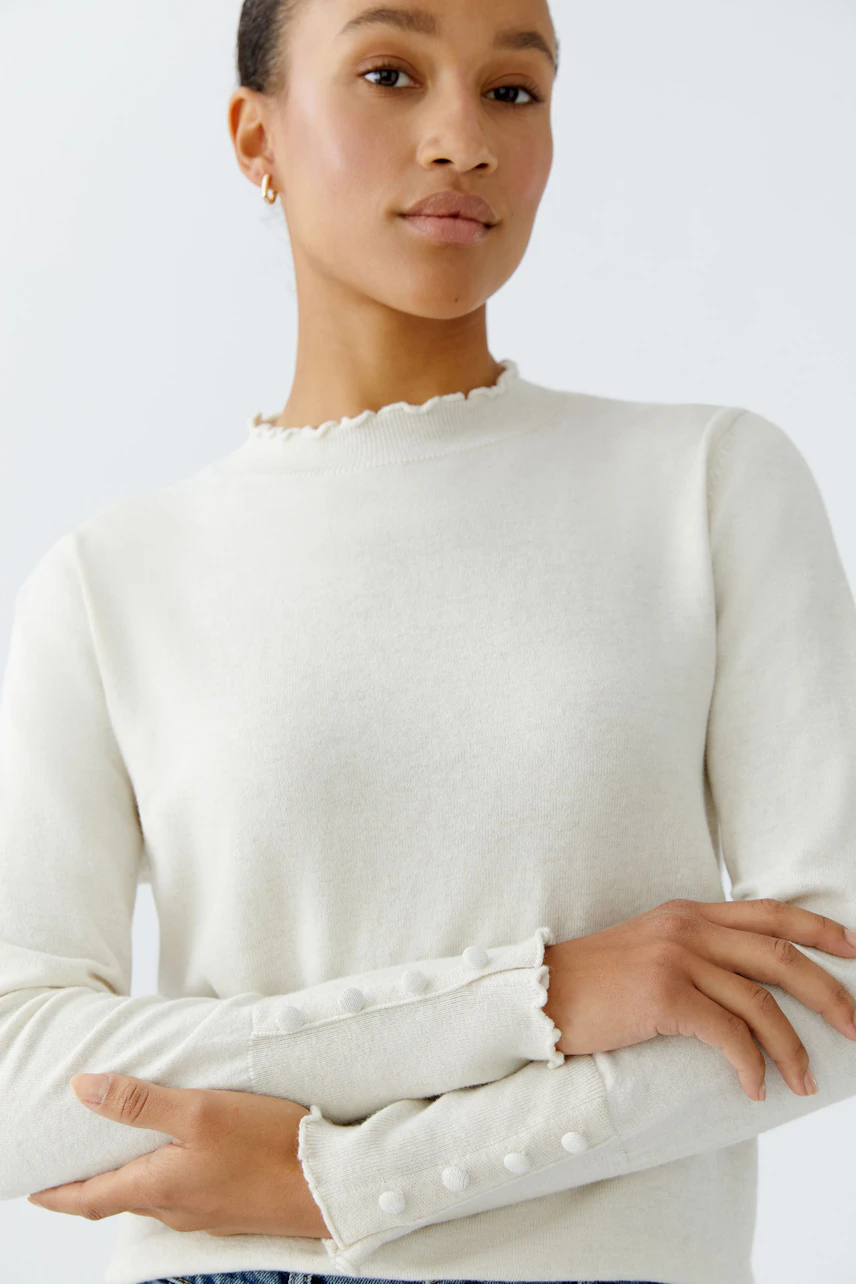 Damen-Pullover: Frau in cremefarbenem Pullover mit Rüschensaum und Knöpfen an den Ärmeln.