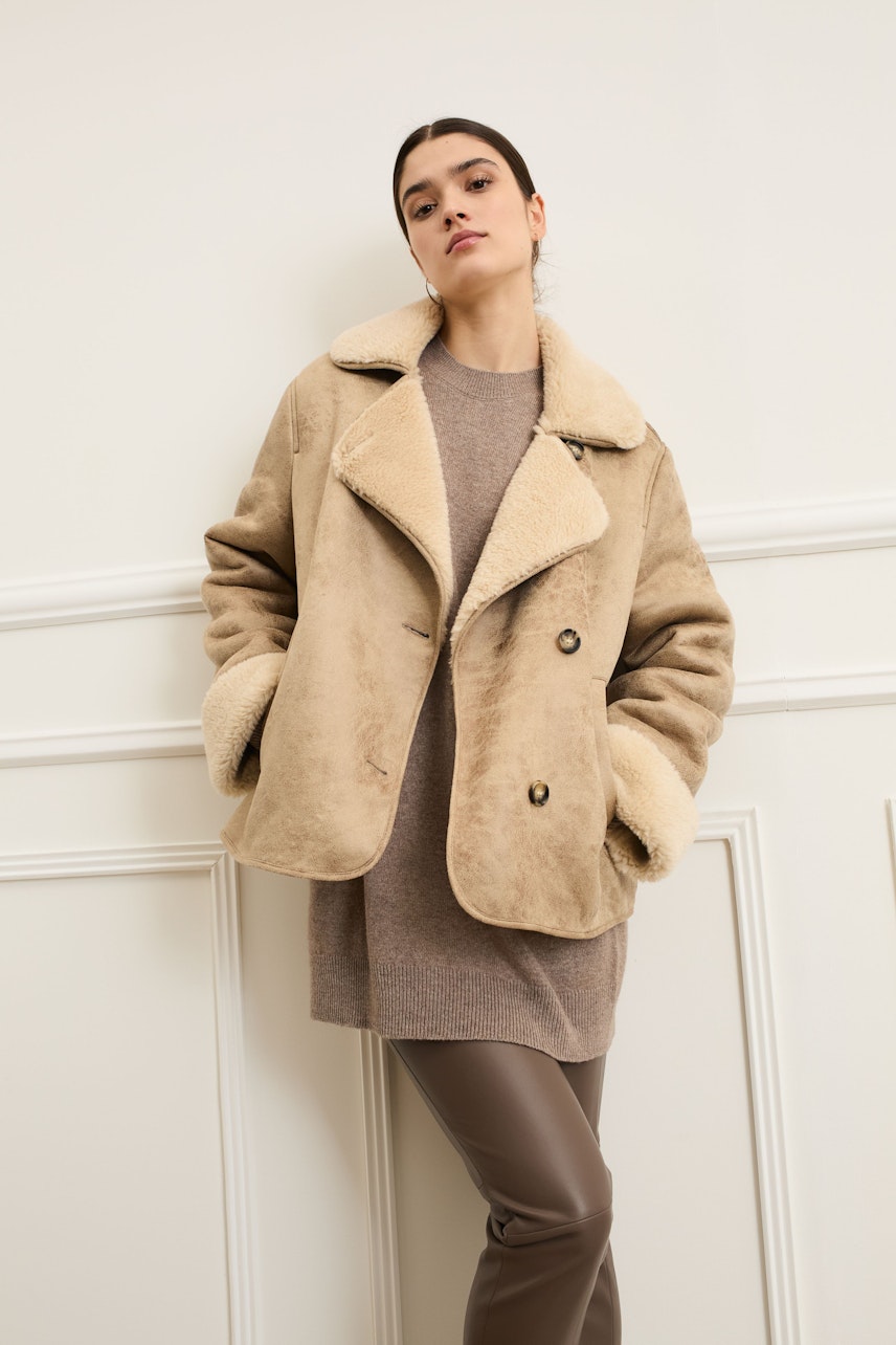 Beige Outdoorjacke: Frau in lässiger Jacke mit weichem Kragen und Knöpfen, kombiniert mit einem grauen Pullover.