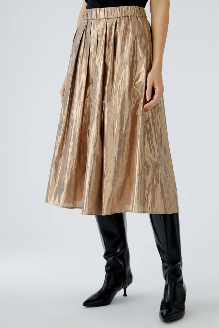 Stylish women's pleated skirt in shiny bronze, paired with knee-high black boots.