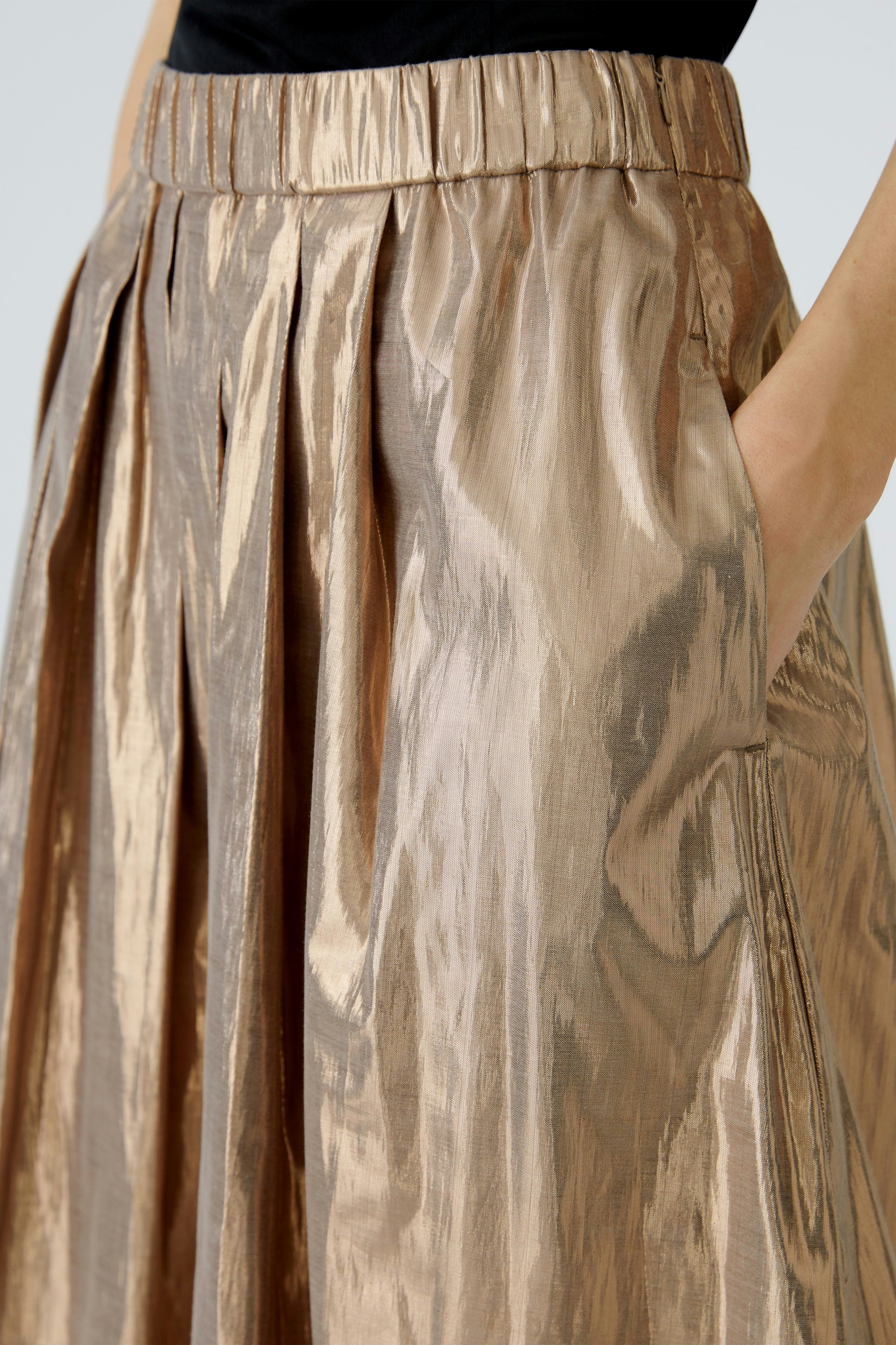 Faltenrock in glänzendem Material: Frau trägt einen beigen Faltenrock mit seitlichen Taschen.