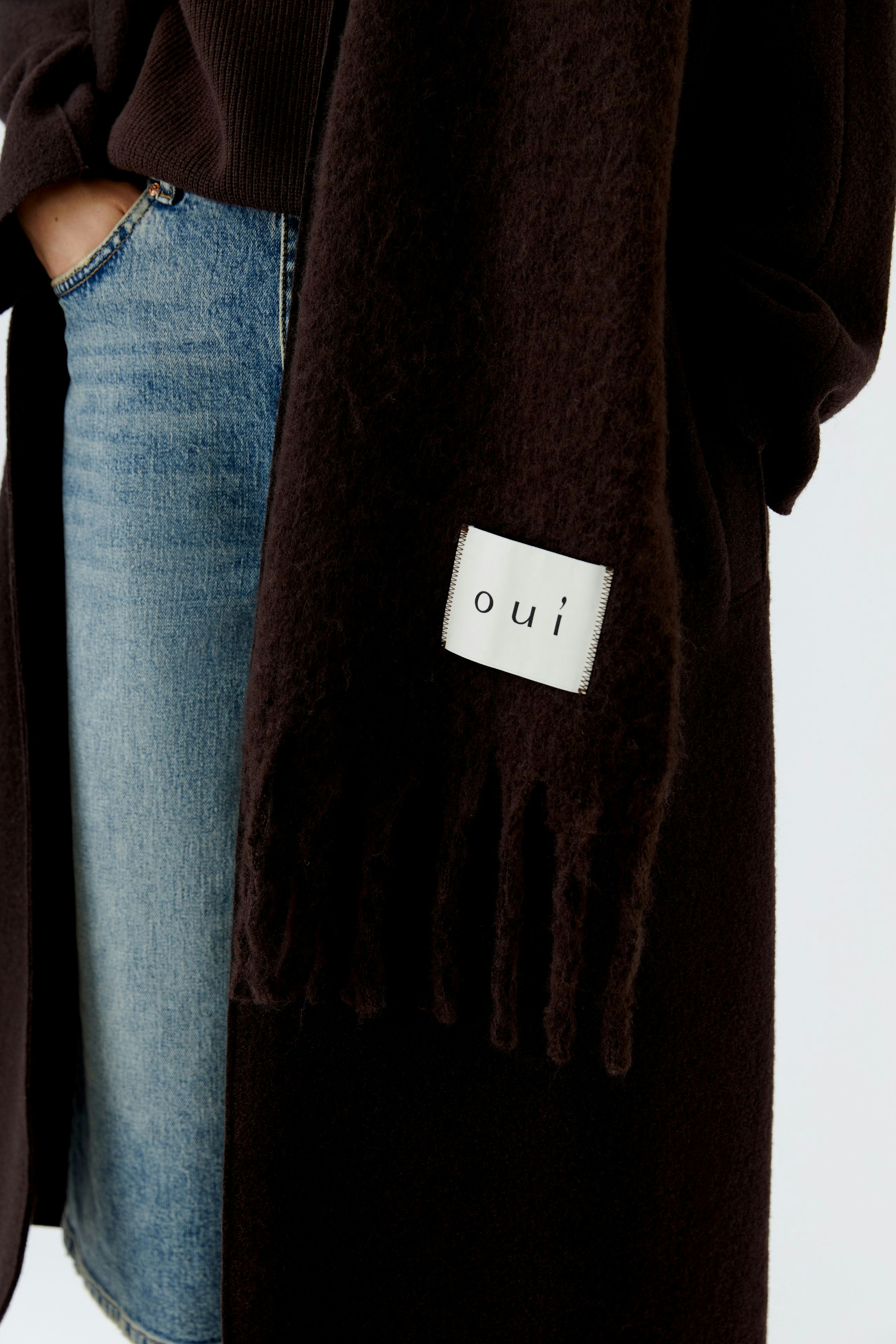 Damen-Schal in Dunkelbraun mit Fransen und Logo-Detail, lässig kombiniert mit Jeans.