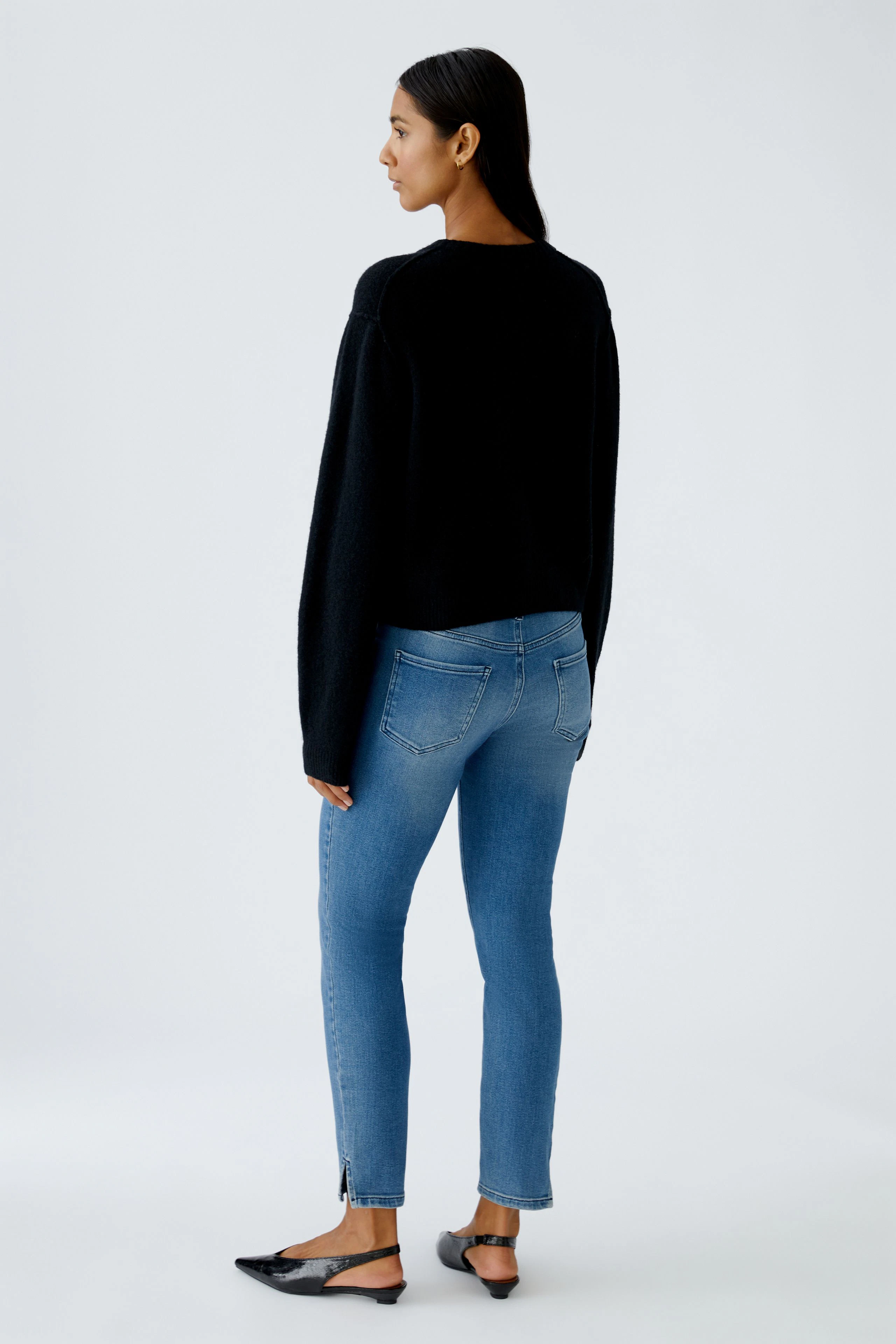 Damen-Strickjacke: Frau in schwarzer Strickjacke und blauen Jeans steht lässig mit dem Rücken zur Kamera.