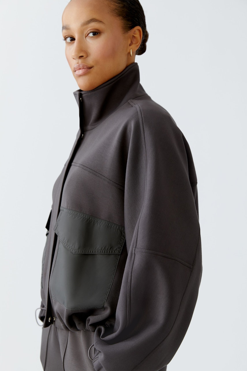 Moderne Damen-Jacke: Frau in grauer Jacke mit hohem Kragen und praktischer Tasche.