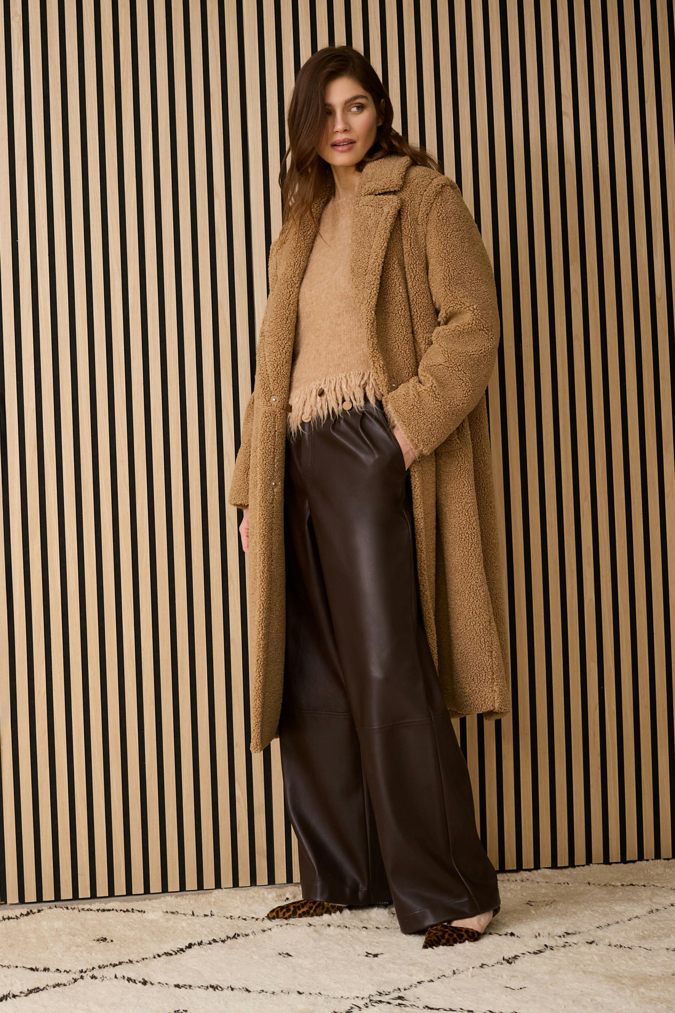 Cozy winter coat: Woman in a soft brown coat over a beige pullover and leather pants, standing confidently.