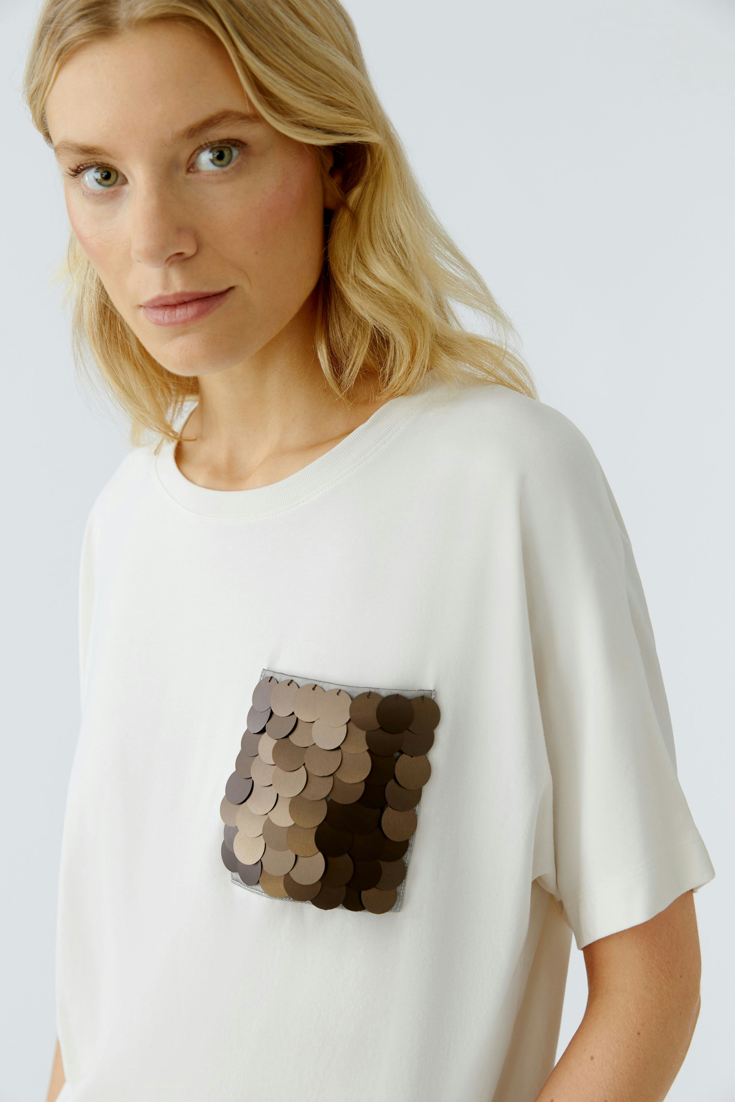 Casual women's T-shirt: Young woman in a white tee with a sequined pocket, looking confidently at the camera.