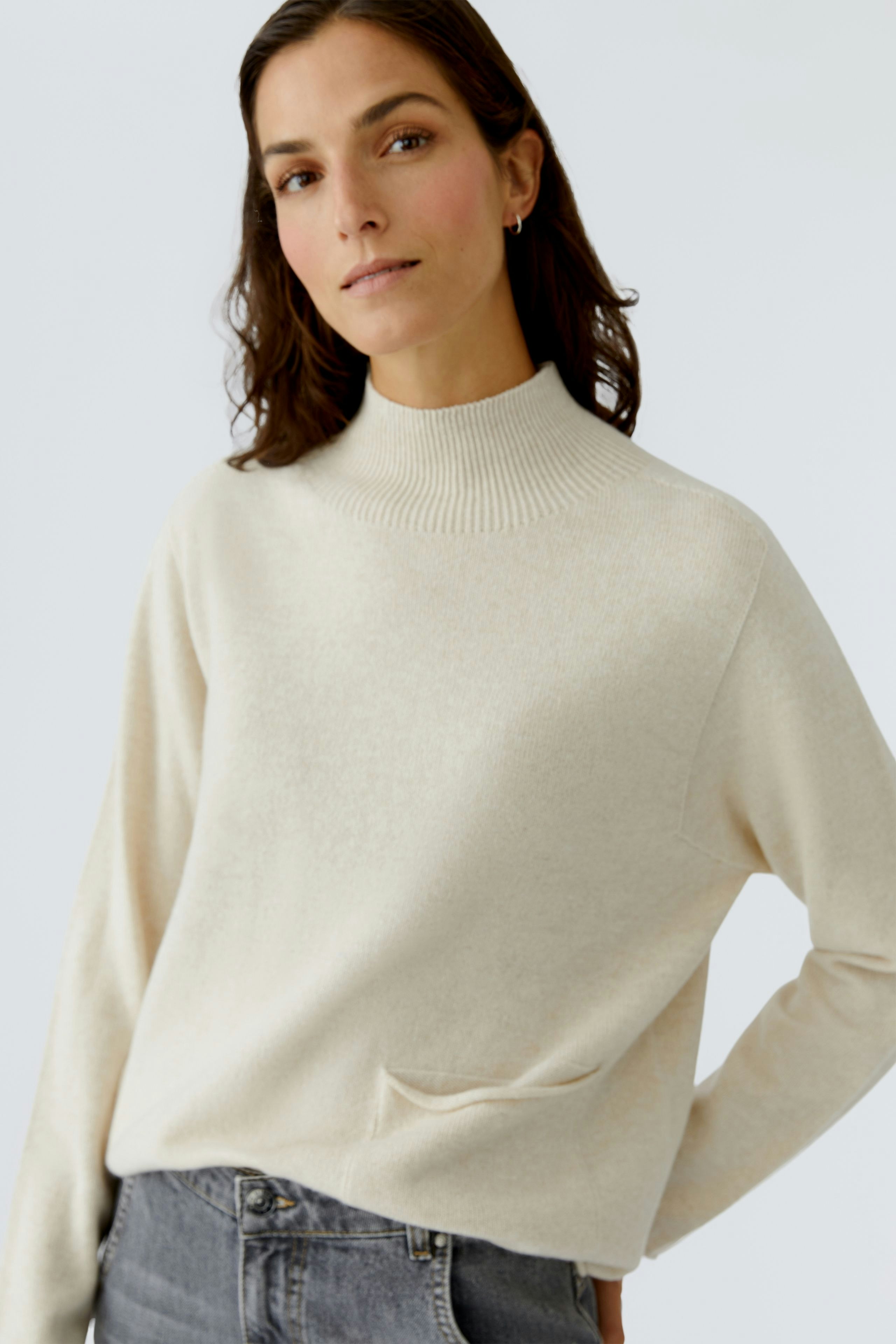 Modern women's pullover: A woman in a cream pullover with a high neck and pocket, looking confidently.