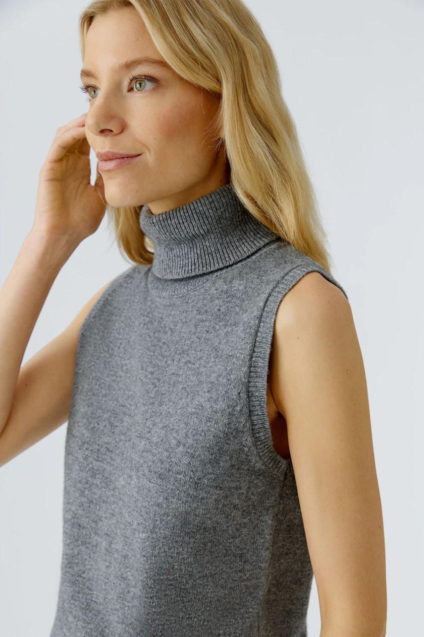Casual women's top: Young woman in a sleeveless gray pullover with a high collar, looking thoughtfully.