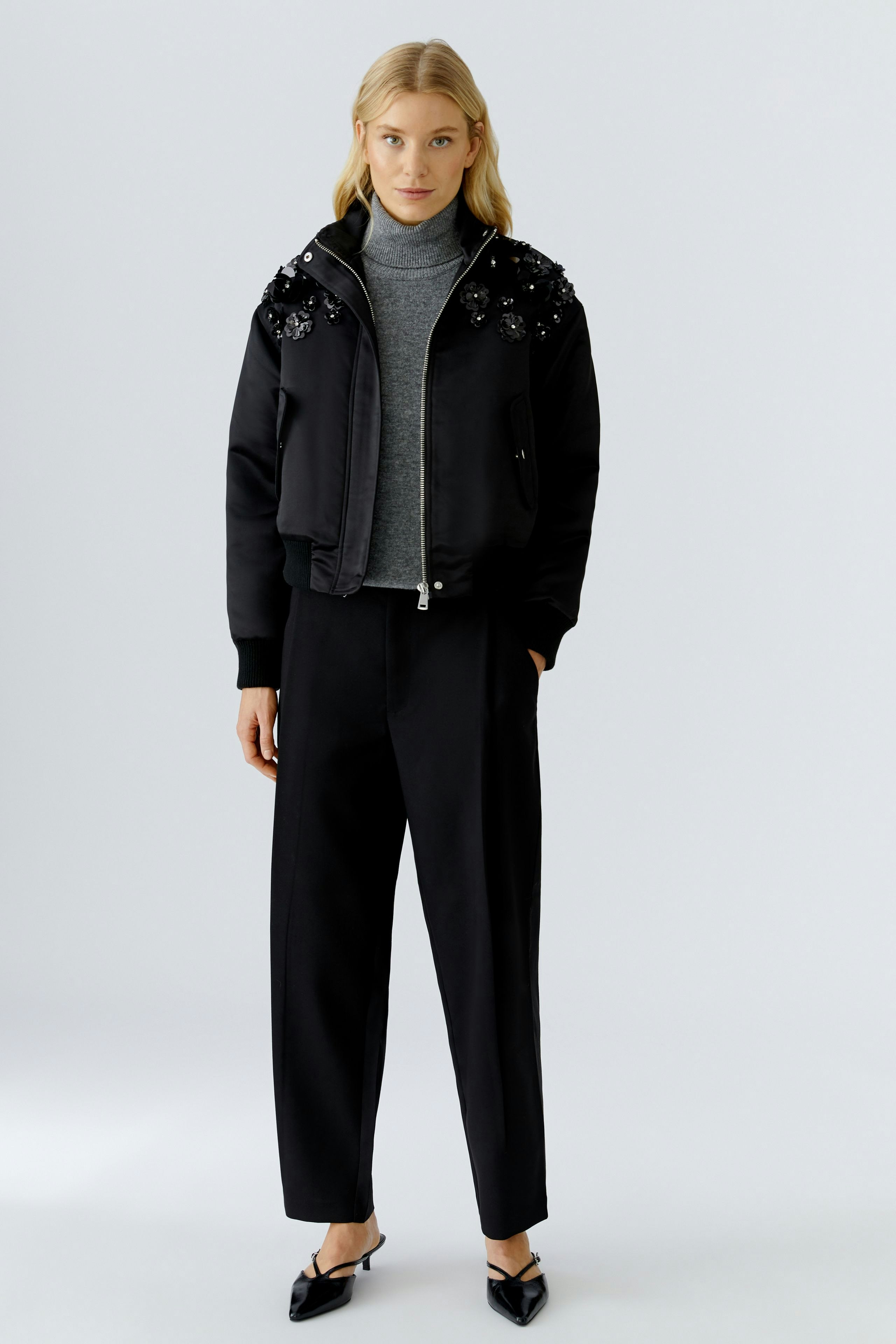 Stylish women's pleated trousers: A woman in black pleated trousers paired with a grey top and bomber jacket.