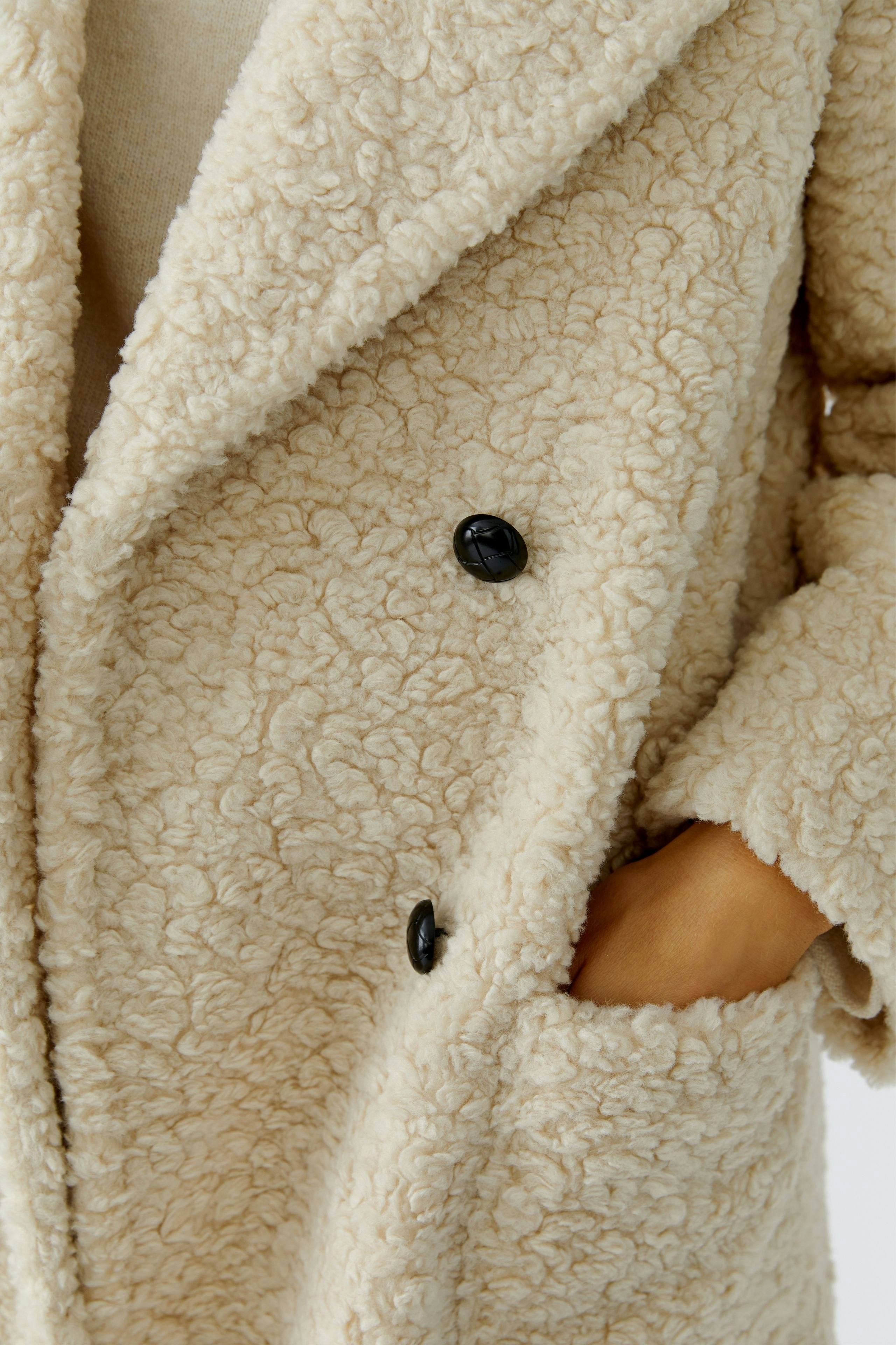 Cozy women's coat: A woman in a soft, textured cream coat with black buttons and pockets.