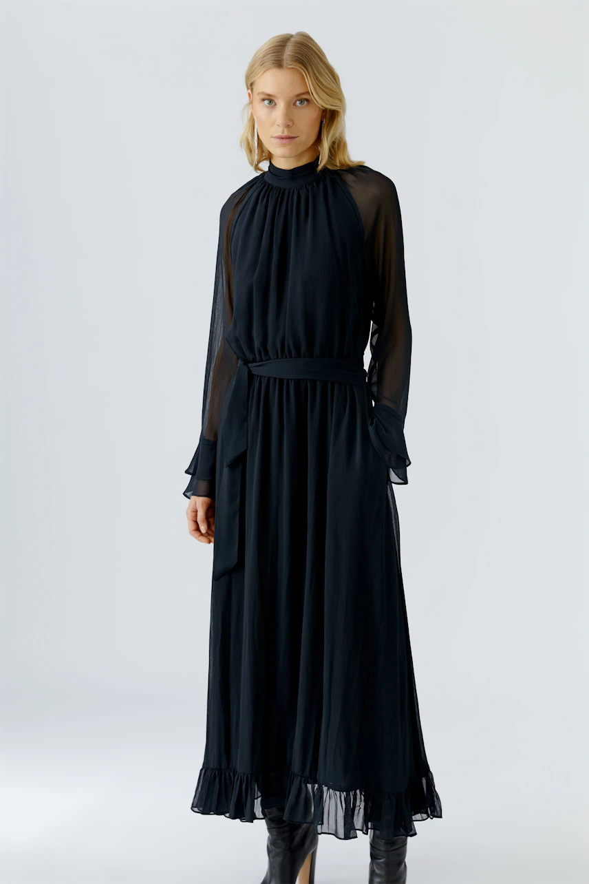 Maxikleid: Frau in schwarzem, fließendem Maxikleid mit transparenten Ärmeln und Rüschensaum.