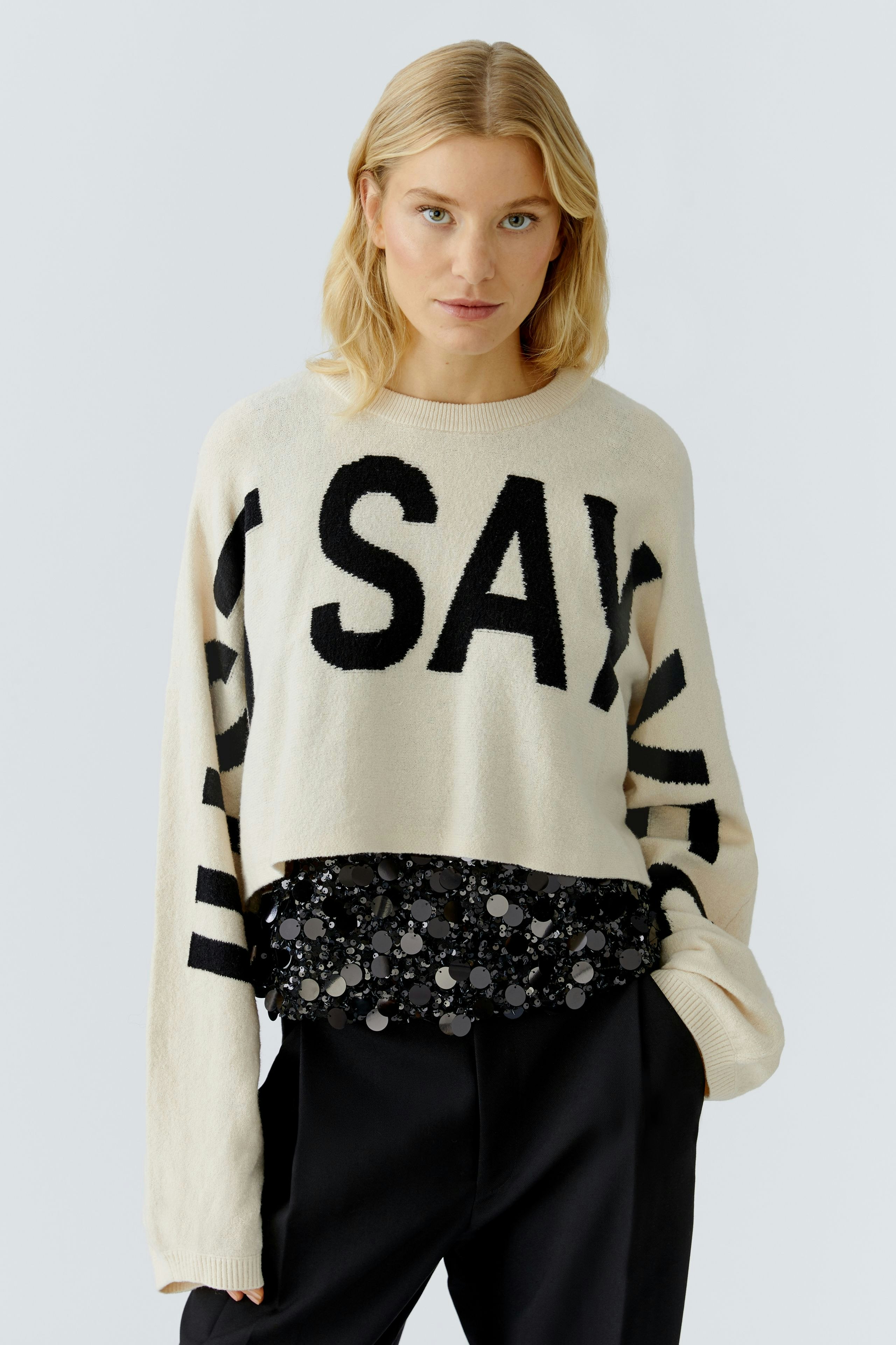 Damen-Pullover: Junge Frau in beigem Pullover mit schwarzem Schriftzug und glitzerndem Saum.