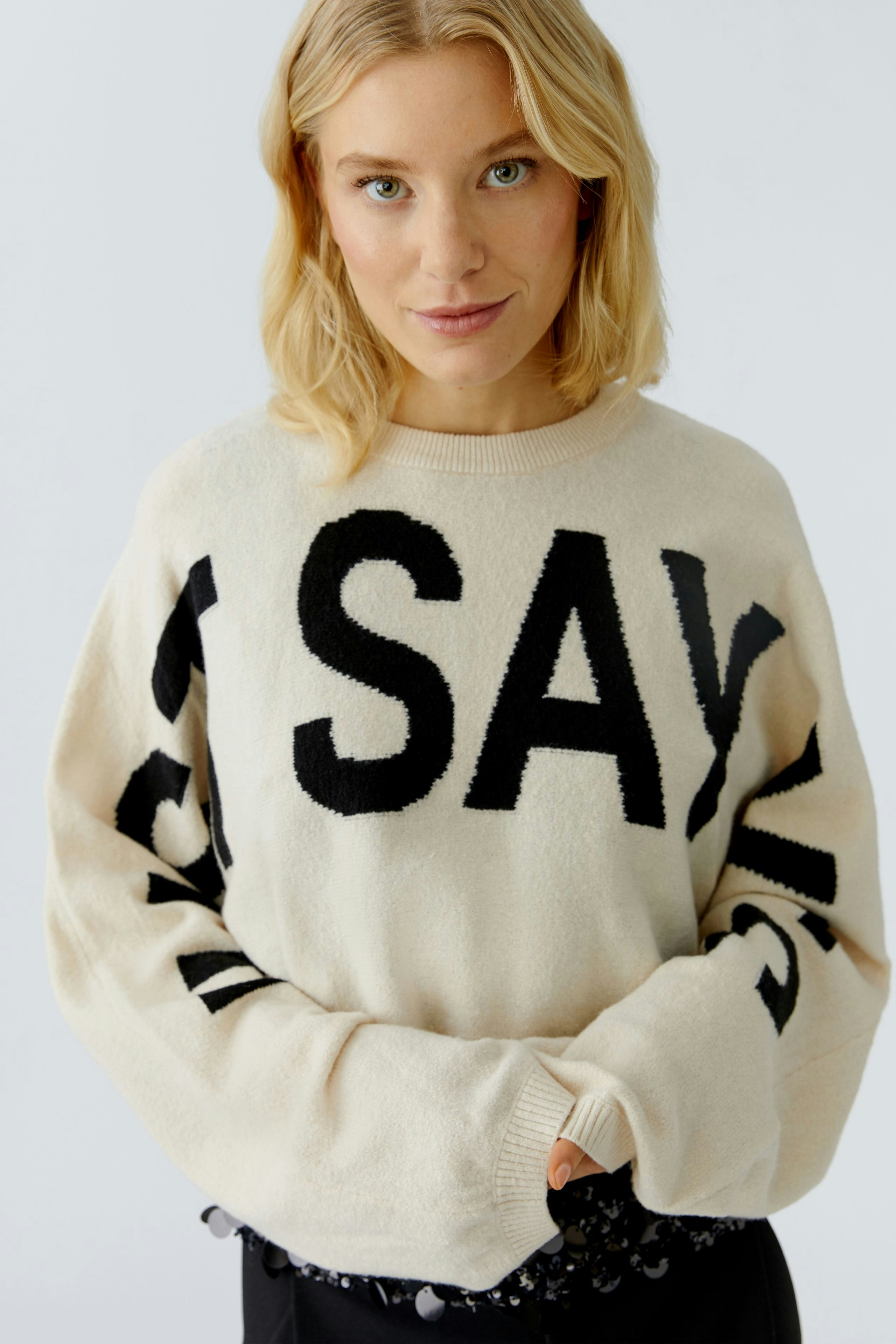 Bequemer Damen-Pullover in Creme mit auffälligem Schriftzug. Frau trägt ihn lässig und selbstbewusst.