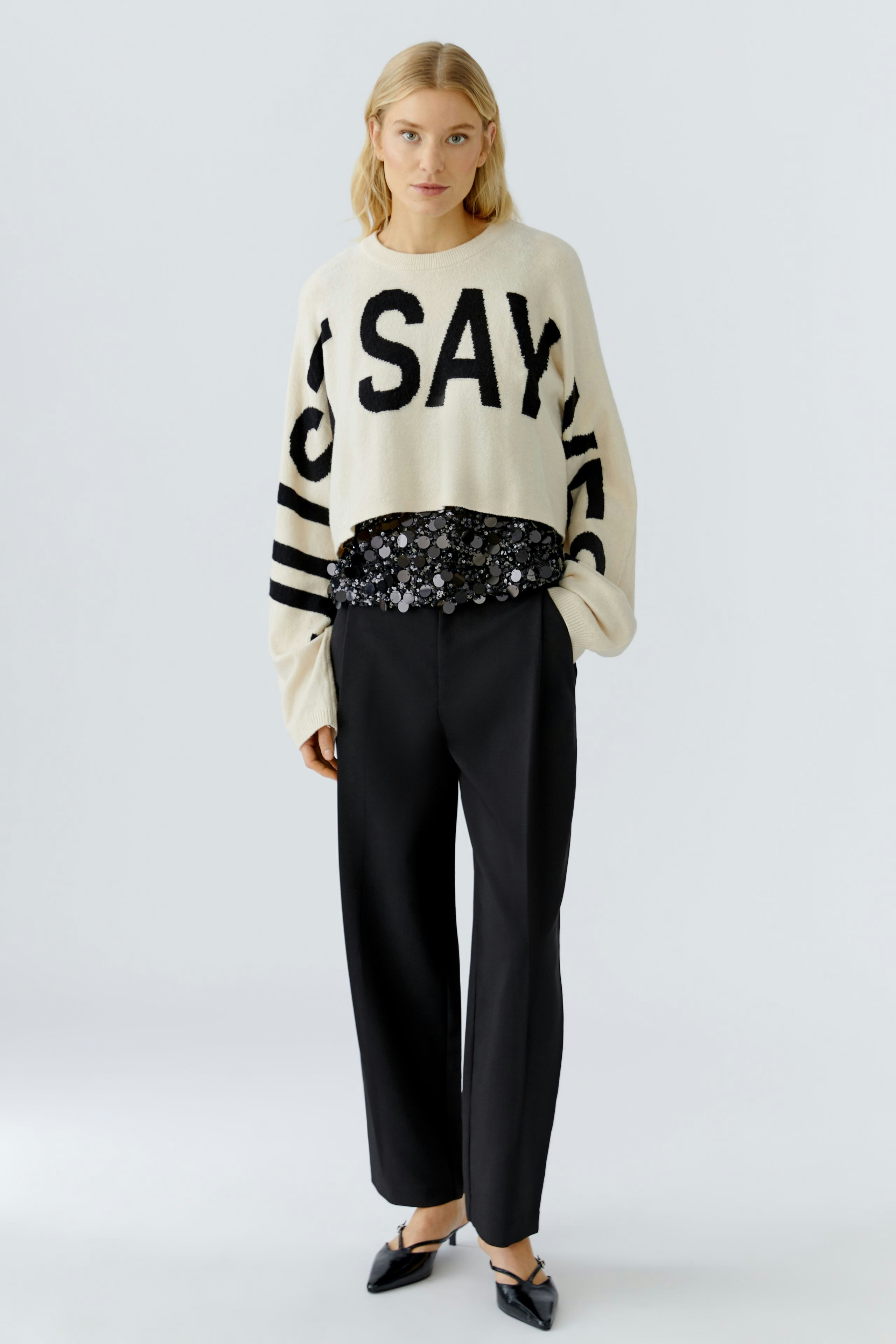 Damen-Pullover: Frau trägt einen beigen Pullover mit schwarzem Schriftzug und lässiger schwarzer Hose.