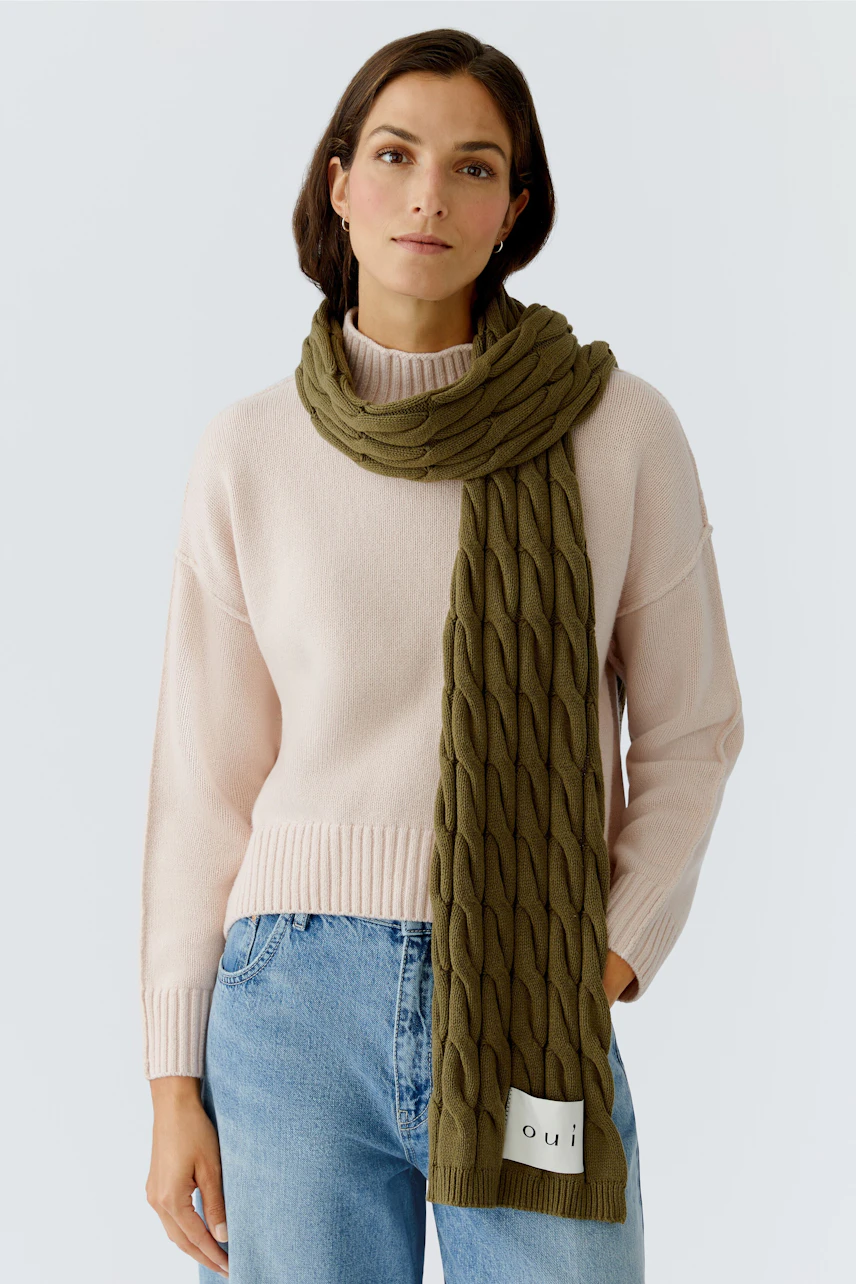 Cozy women's scarf: A woman wearing a green knitted scarf over a light pink pullover, looking relaxed.