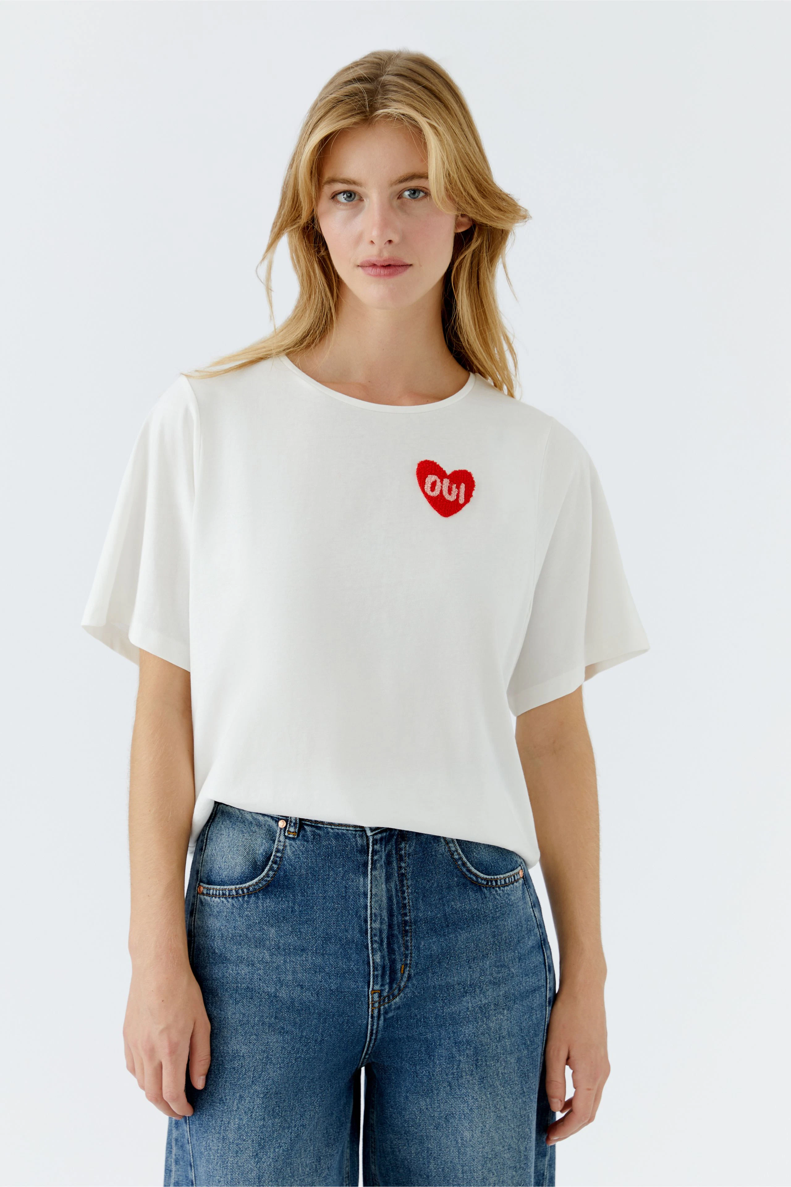 Casual women's T-shirt: Young woman in a white tee with a red heart and 'OUI' logo, paired with blue jeans.