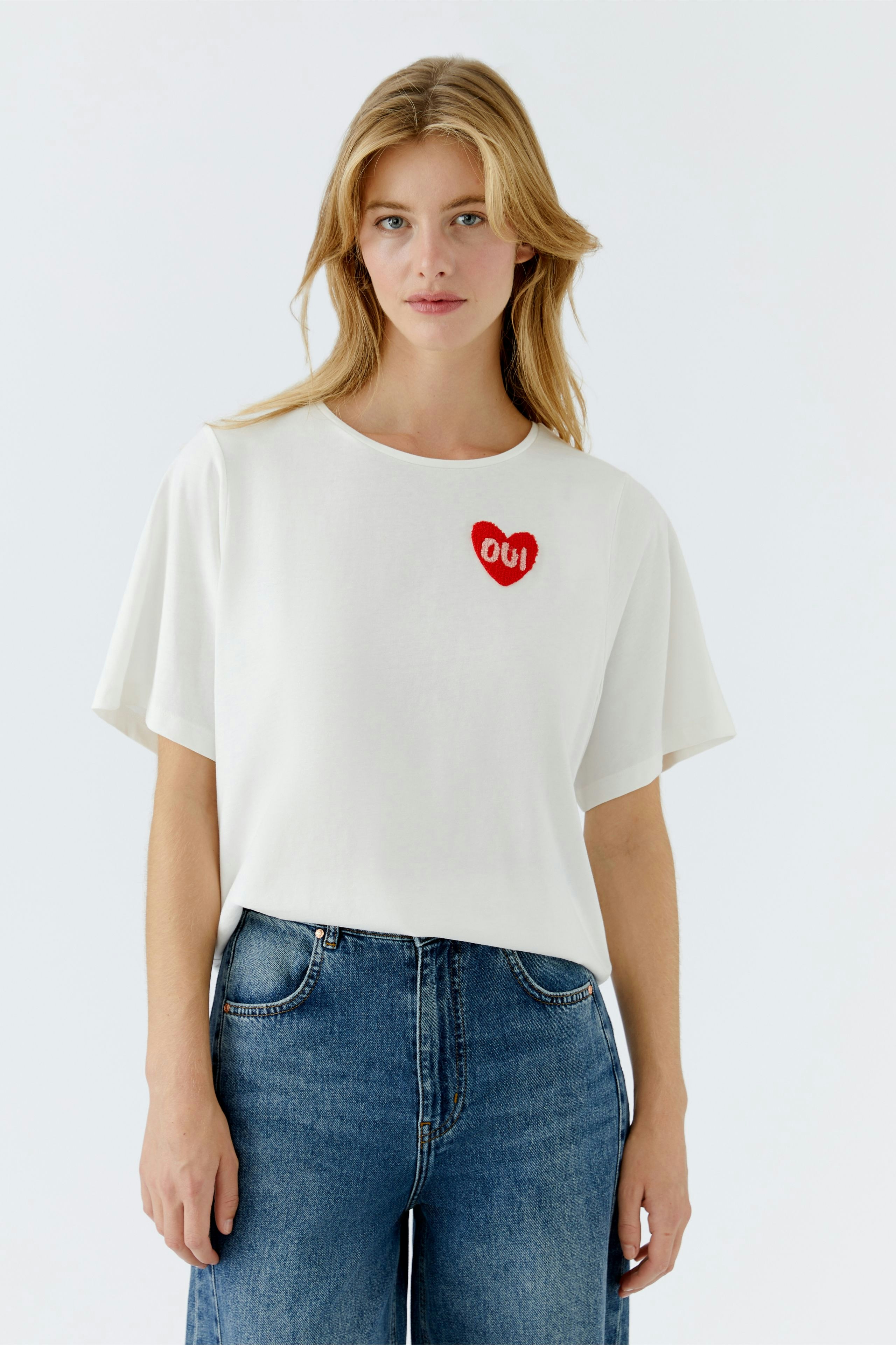 Casual women's T-shirt: Young woman in a white tee with a red heart and 'OUI' logo, paired with blue jeans.