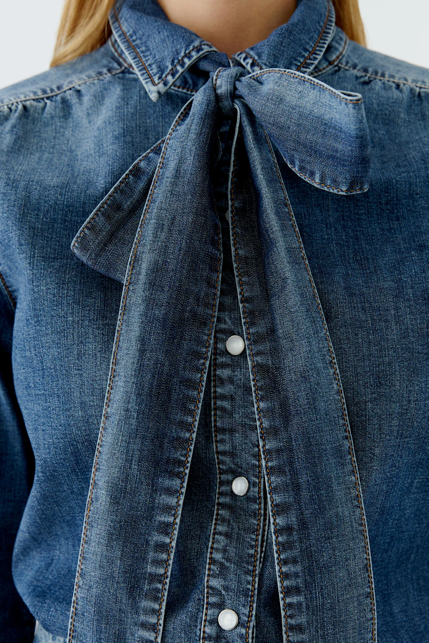 Damen-Jeanshemd: Frau trägt ein blaues Jeanshemd mit Schleifenkragen und Knopfleiste.