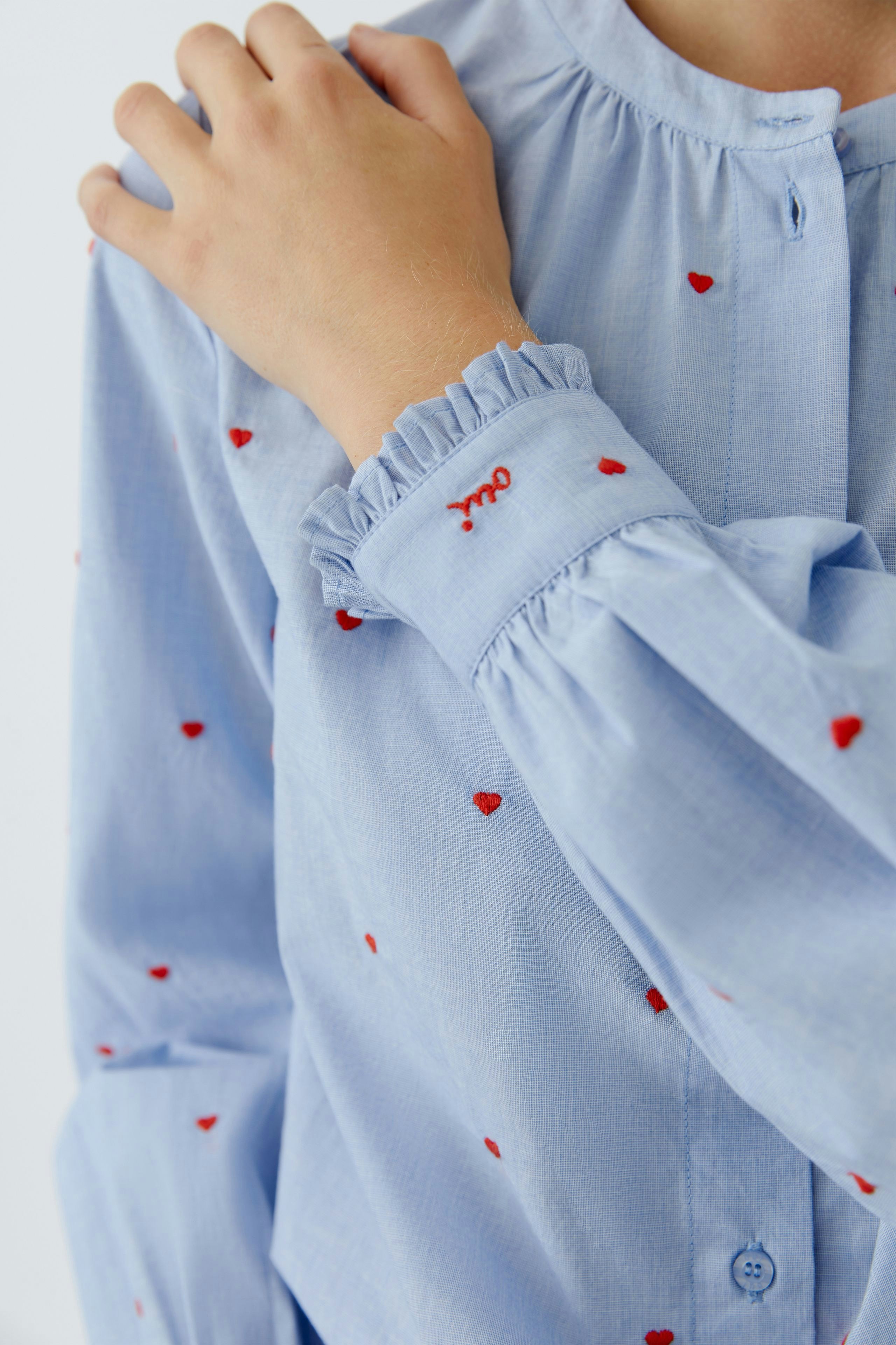 Light blue blouse with red heart details, worn by a woman, featuring a ruffled cuff.