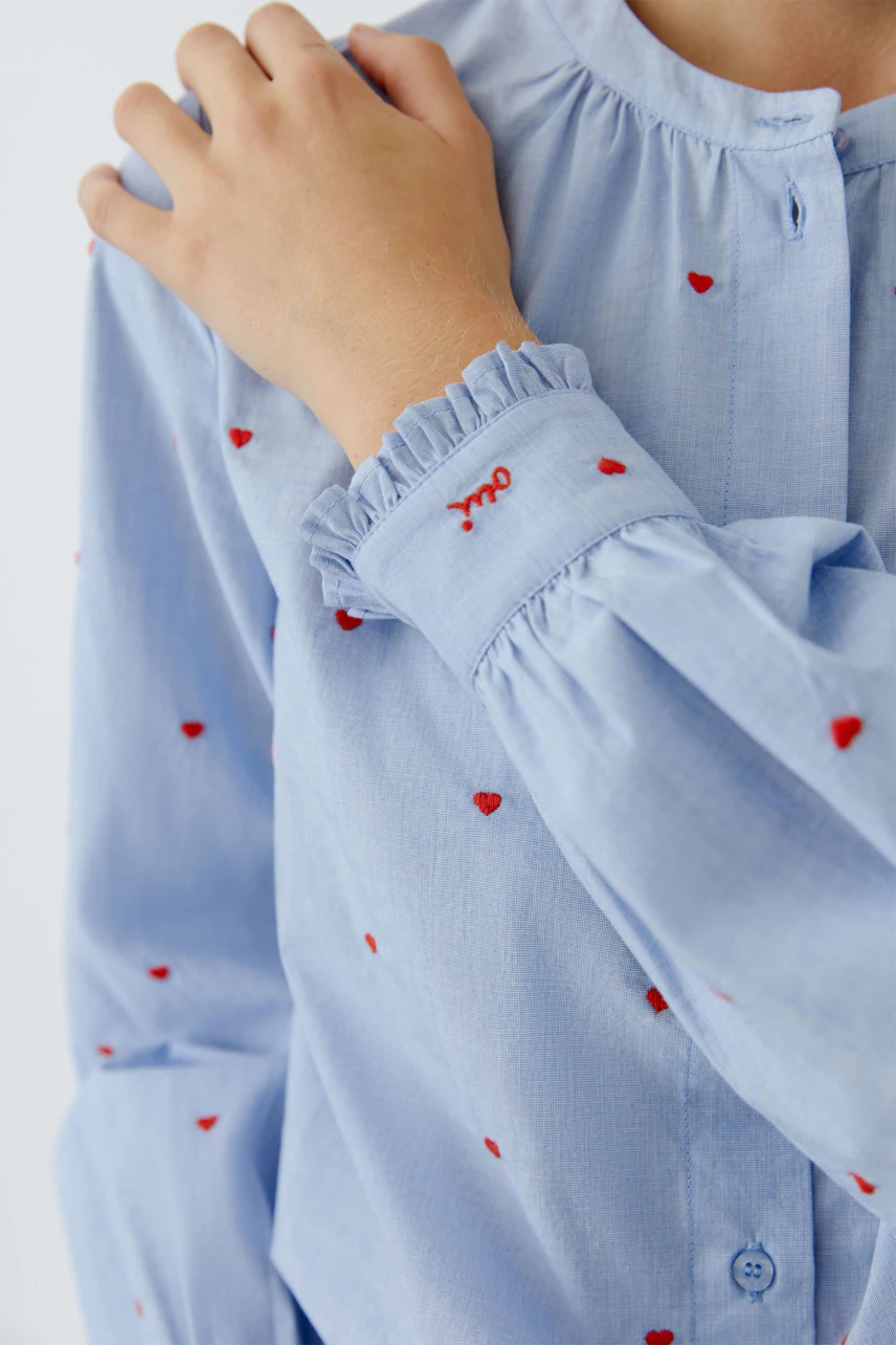 Light blue blouse with red heart details, worn by a woman, featuring a ruffled cuff.