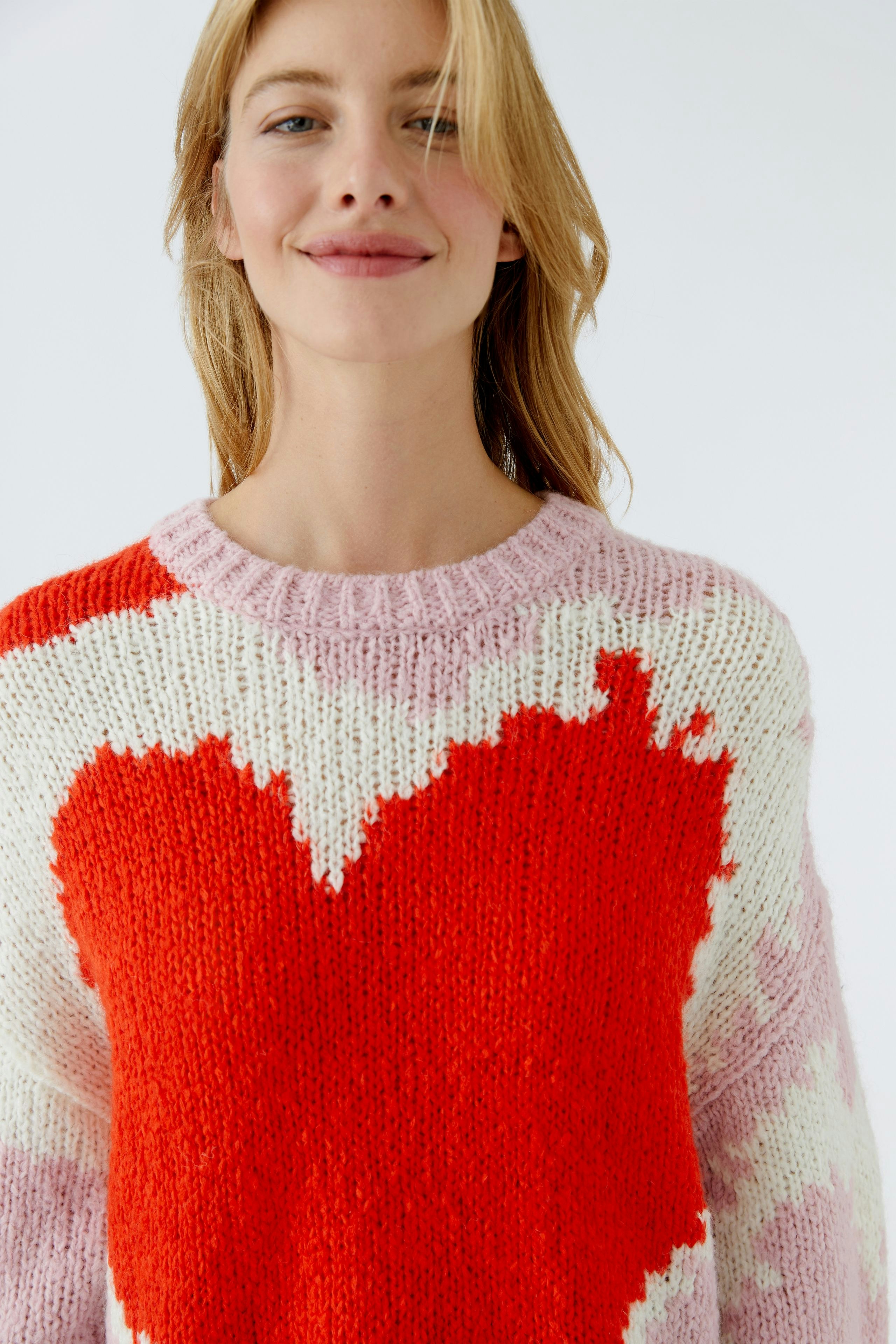 Bunter Damen-Pullover: Frau trägt einen grob gestrickten Pullover mit Herz-Muster in Rot und Rosa.
